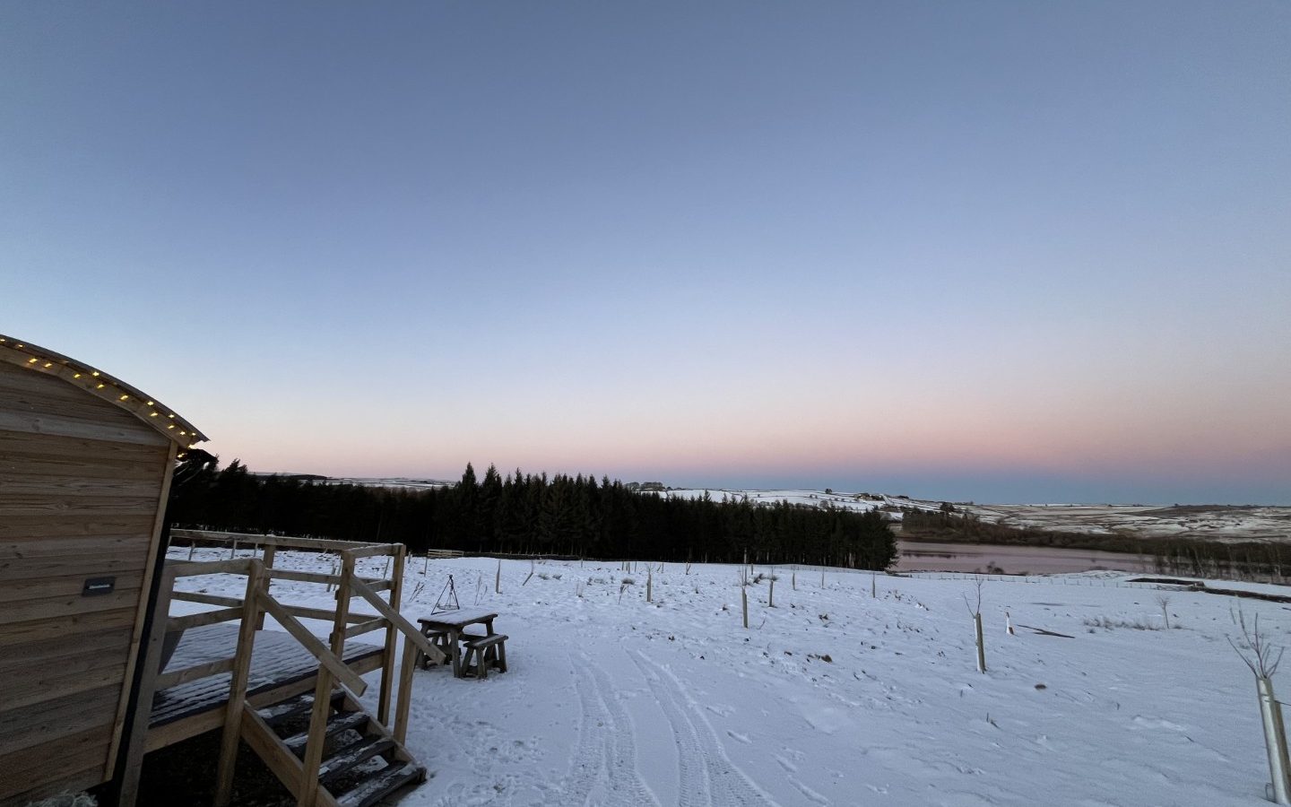 Snow-covered landscape with a wooden cabin at Breaks Fold Farm, a picnic table, and distant hills at dawn or dusk.