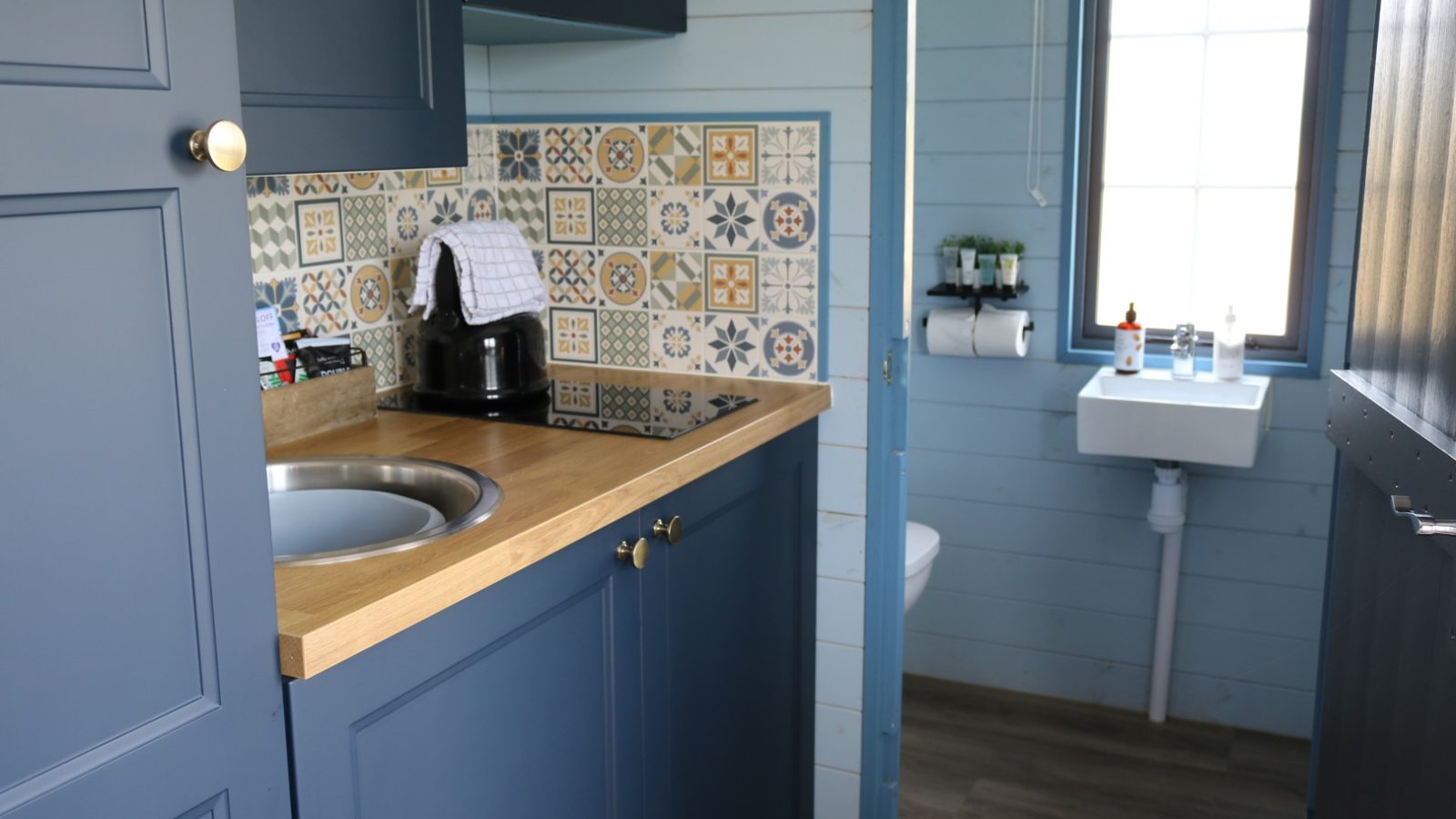 Compact kitchen and bathroom area with blue cabinetry, tile backsplash, sink, small countertop, window, and farmhouse charm.