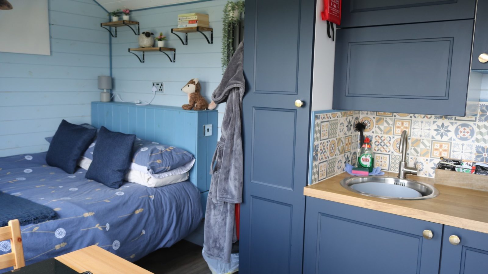 Compact cabin interior with a bed, shelves, and a kitchenette featuring blue cabinets and a colorful tiled backsplash—perfect for breaks on the Fold Farm.