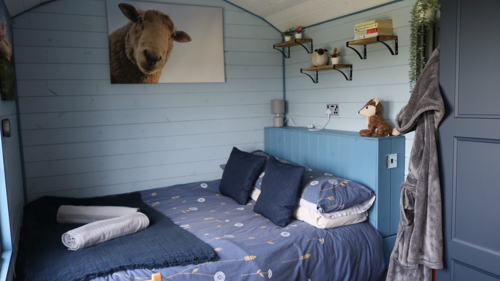 Cozy bedroom at Breaks Fold Farm with blue wooden walls, double bed, sheep artwork, plush toy, and a robe on the door.