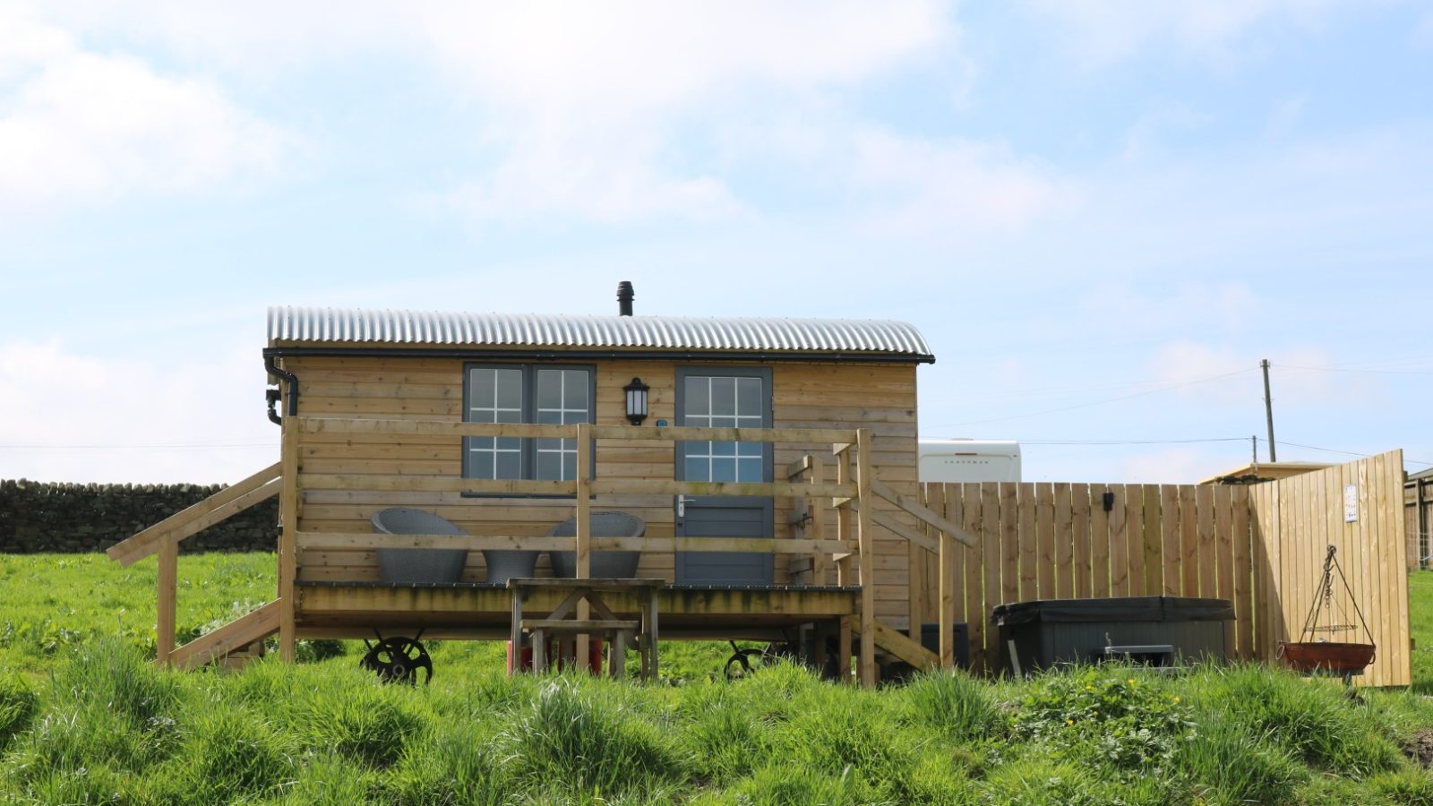 A small wooden cabin with a deck and chairs, set on wheels, rests at Breaks Fold Farm under a partly cloudy sky.