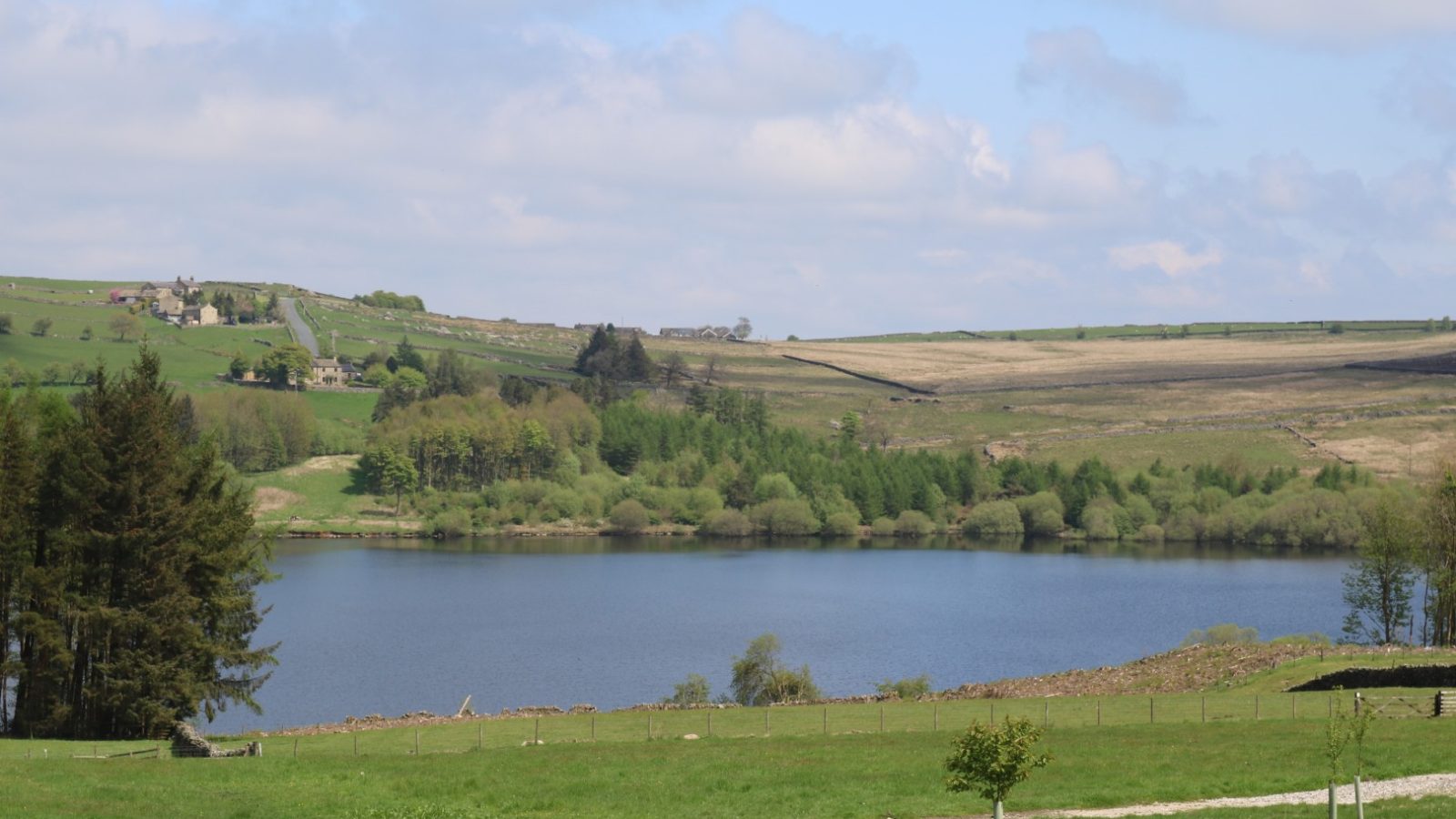 View of serene Breaks Fold Farm, with a lake surrounded by green fields, trees, and hills under a partly cloudy sky.