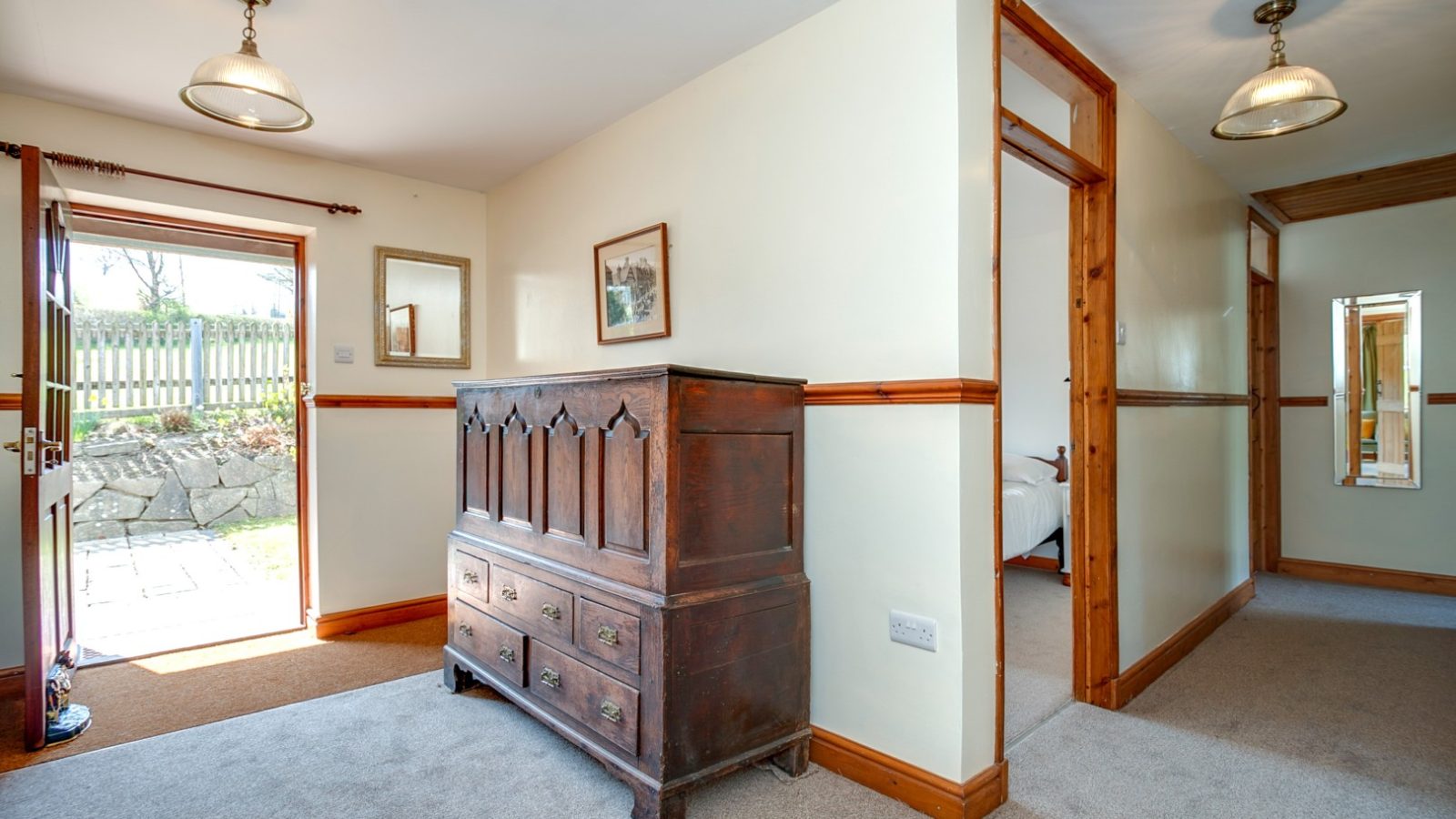 Hallway in Brook Cottage with beige walls, wooden trim, a dark dresser, an open door, hanging lights, and framed wall art.