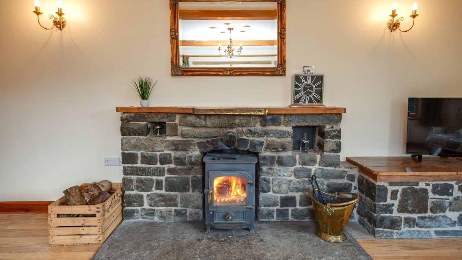 Cozy living room at Brook Cottage with a lit wood-burning stove, stone fireplace, wall mirror, and wooden accents.