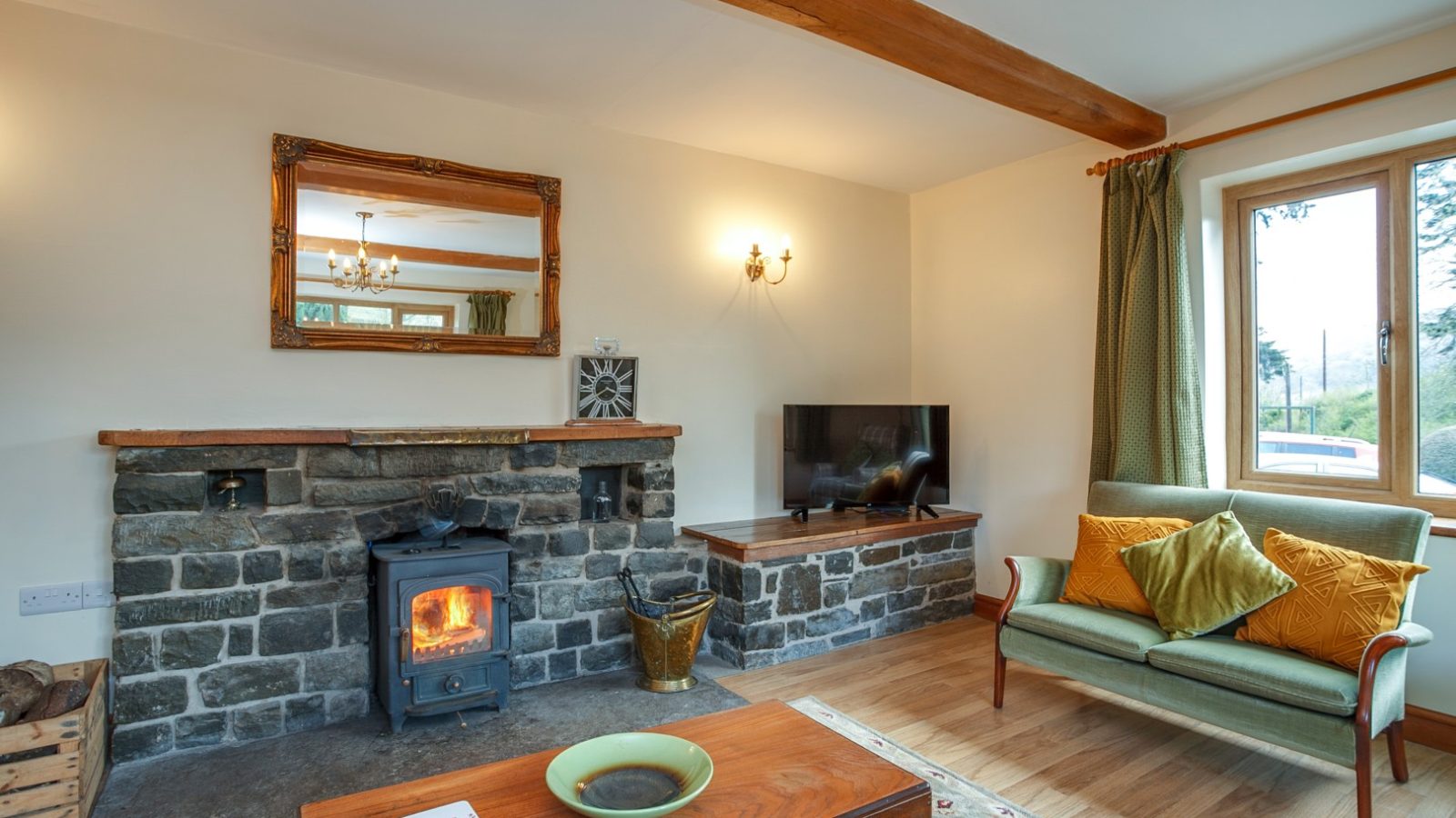 Cozy living room in Brook Cottage with a wood-burning stove, stone fireplace, green armchair, TV, and brown-curtained window.