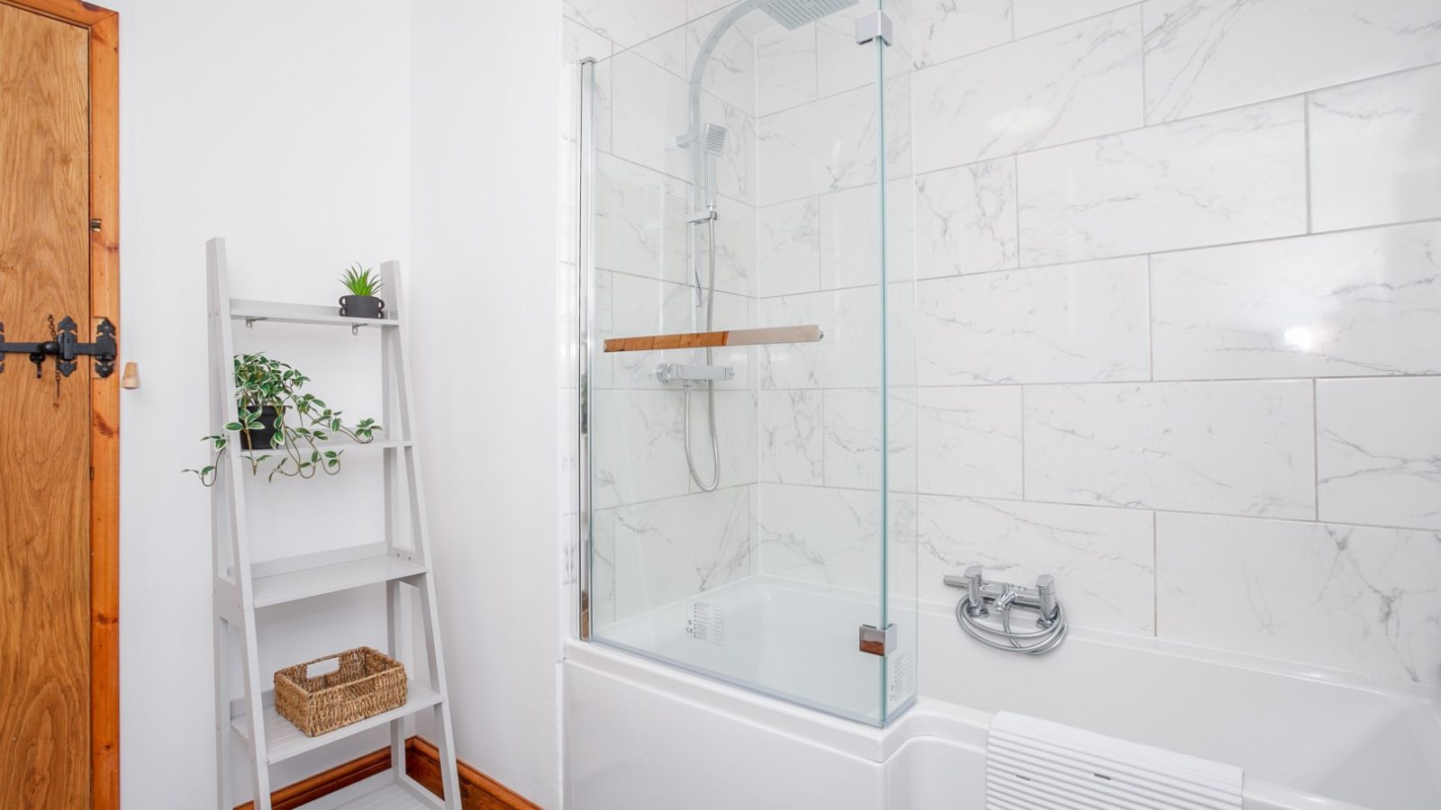 Bathroom at Brook Cottage with a white bathtub, glass shower, marble tiles, wooden shelf of plants, and a wicker basket.