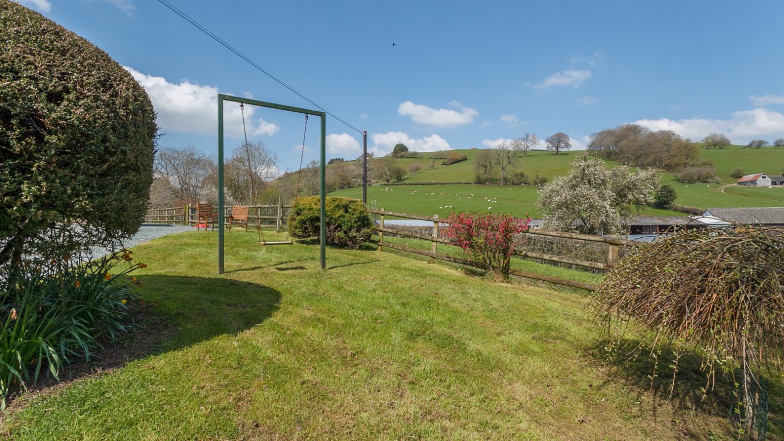 A rural scene with a swing frame on grass near Brook Cottage, surrounded by trees with hills and scattered sheep in the background.