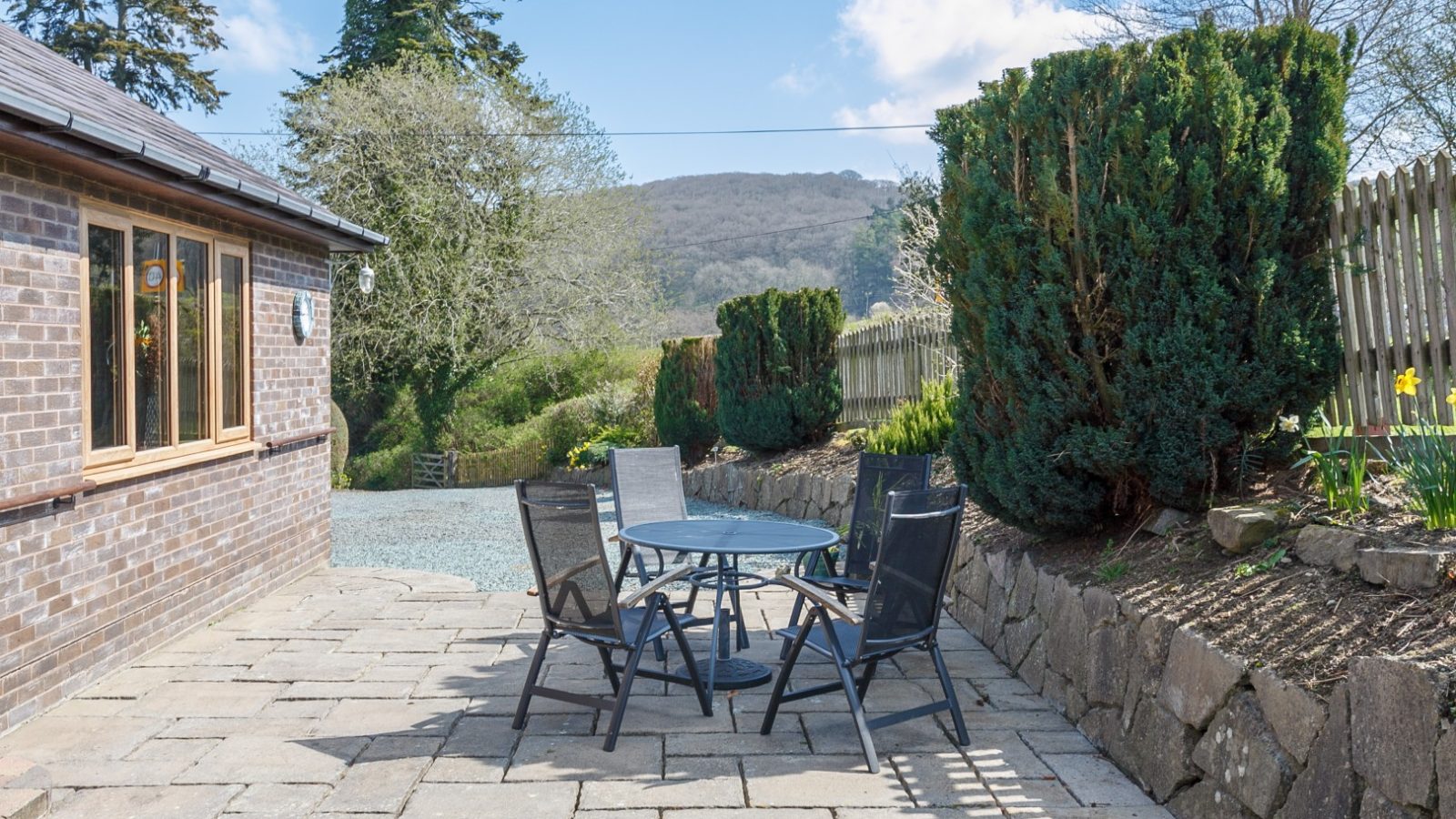 The patio at Brook Cottage, with a glass table and chairs, overlooks a garden nestled against scenic hills.