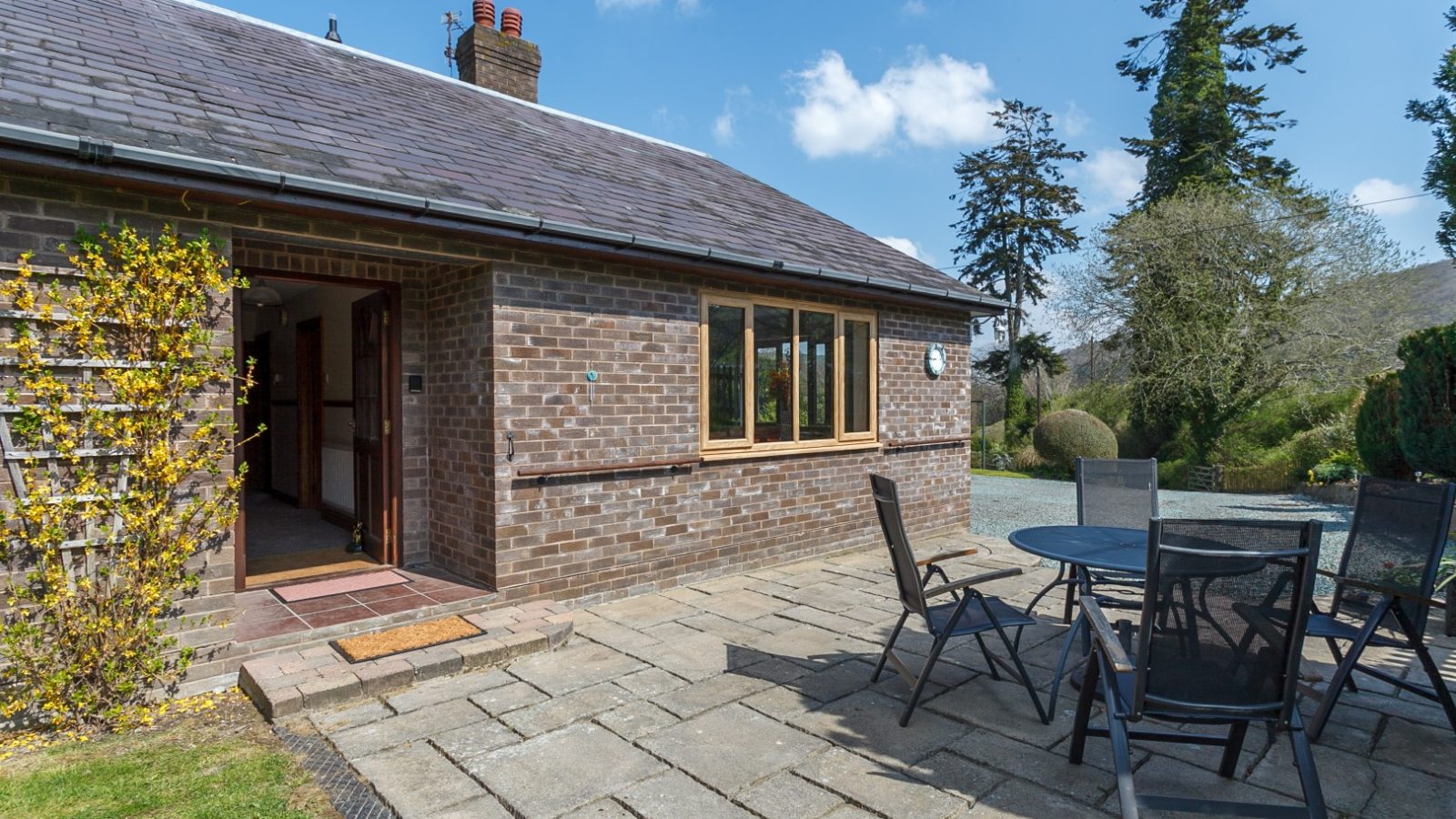 Brook Cottage: A single-story brick house with an open door, patio, round table, and chairs nestled among trees under a blue sky.