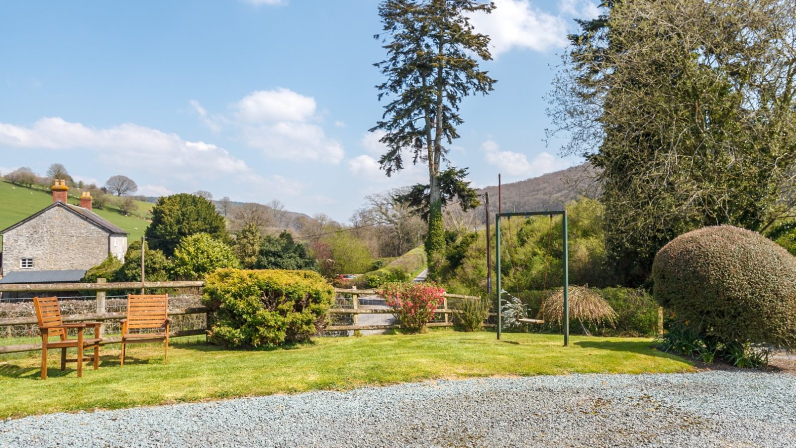 Brook Cottage garden with wooden benches, lush greenery, and distant hills under a blue sky with scattered clouds.