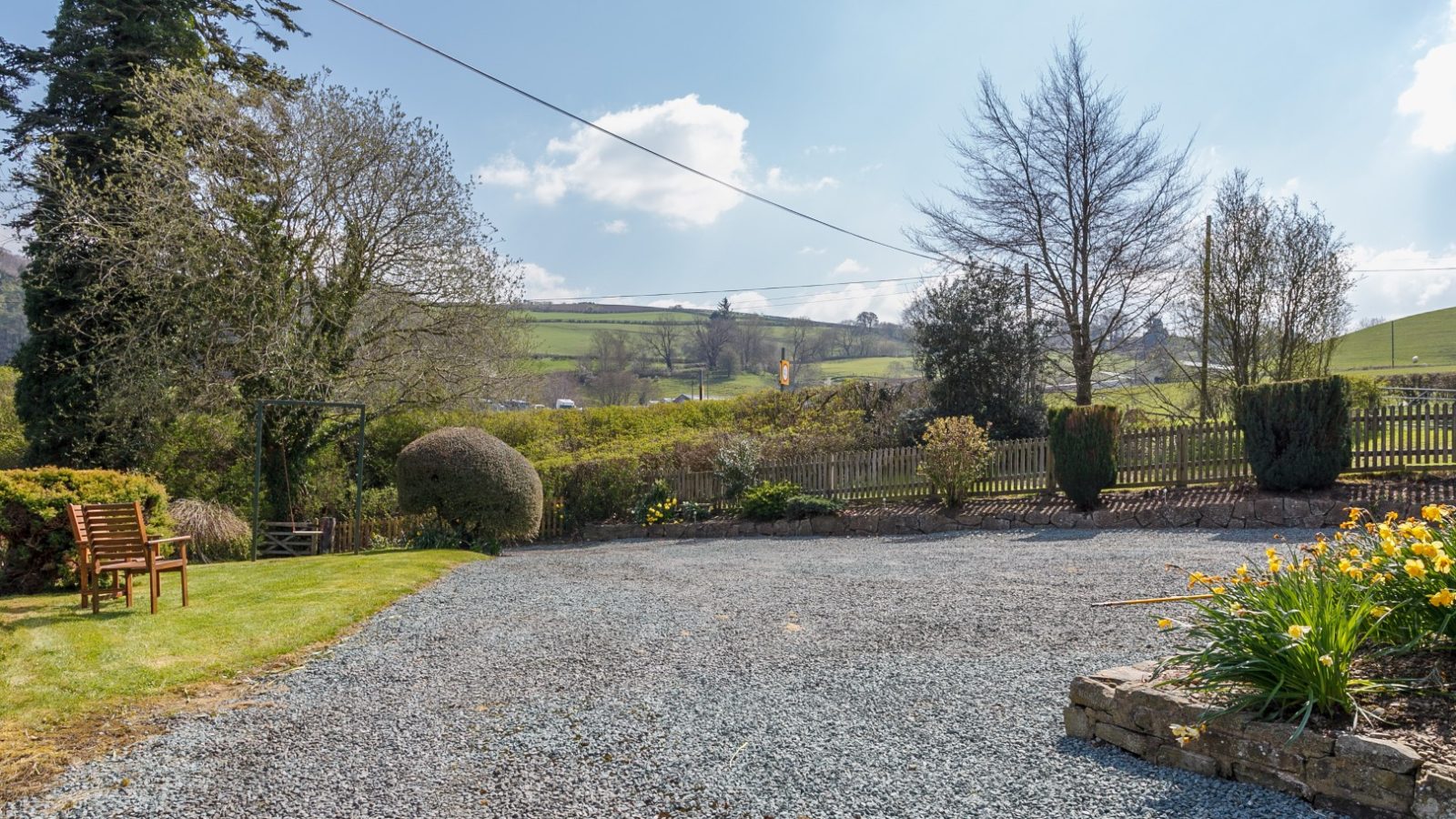 Gravel driveway at Brook Cottage features a wooden bench on grass, daffodils upfront, with a countryside landscape backdrop.