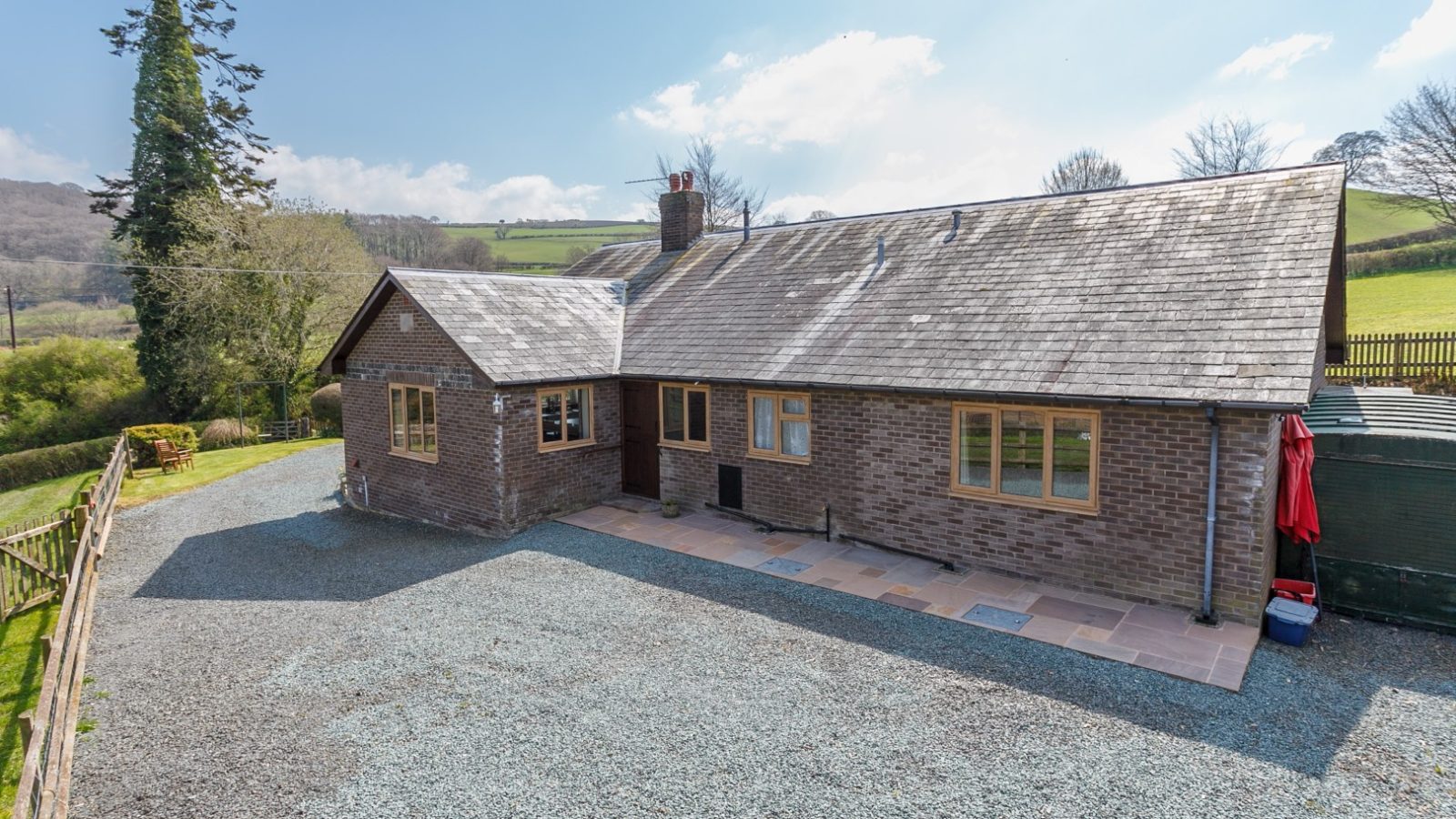 Brook Cottage is a single-story brick house with a slate roof, framed by a gravel driveway and scenic countryside.