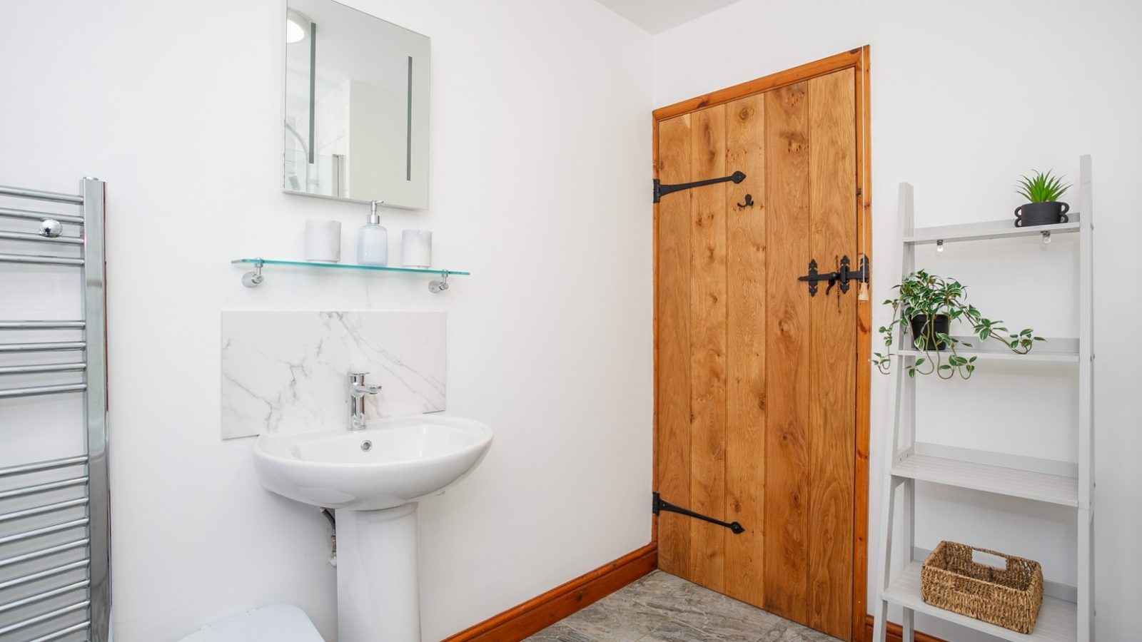 A bathroom at Brook Cottage with a pedestal sink, wooden door, towel rack, mirror, and a white shelf with decor items.