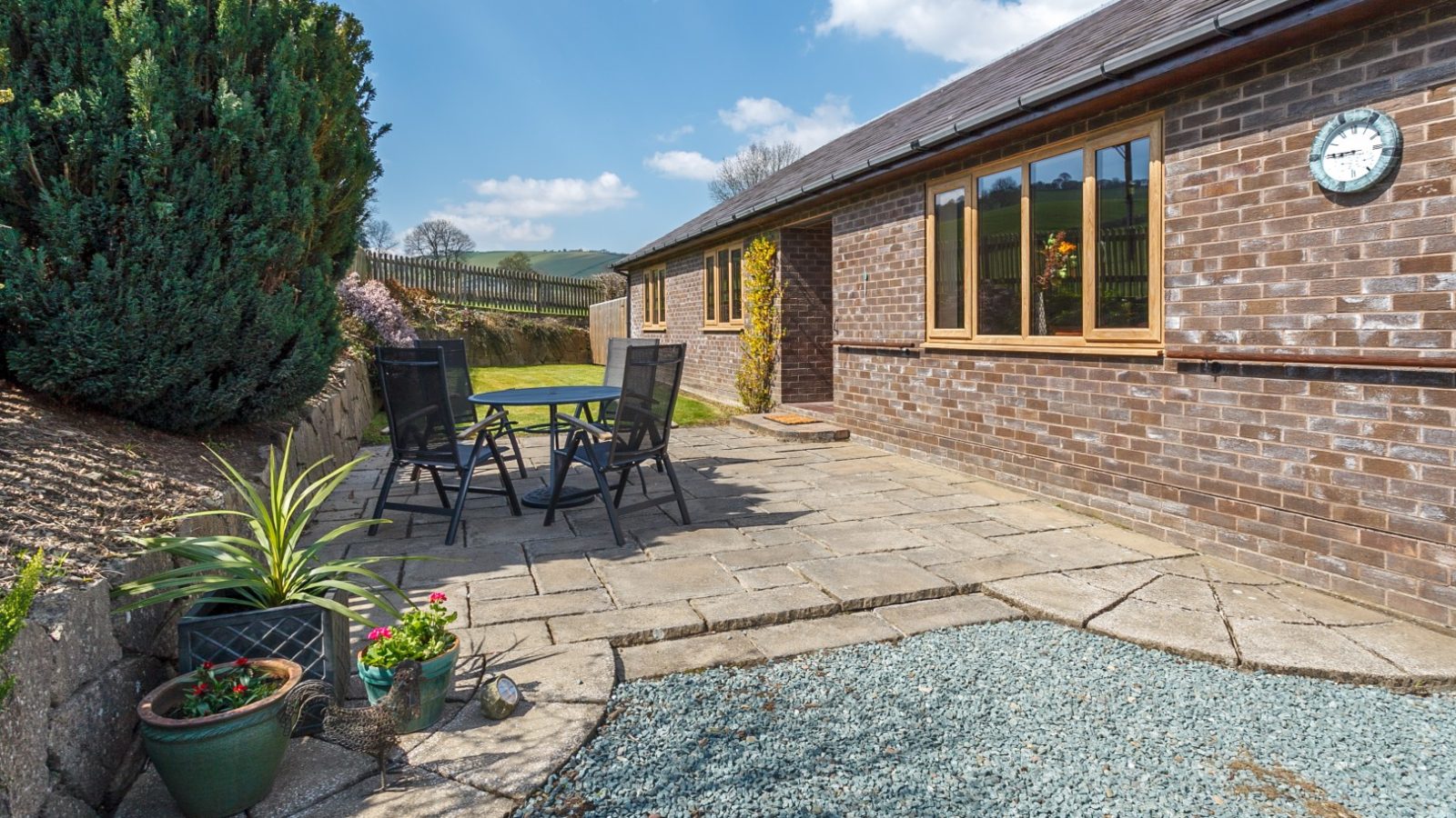 Stone patio at Brook Cottage, featuring a table and chairs, surrounded by shrubs and potted plants under a sunny blue sky.