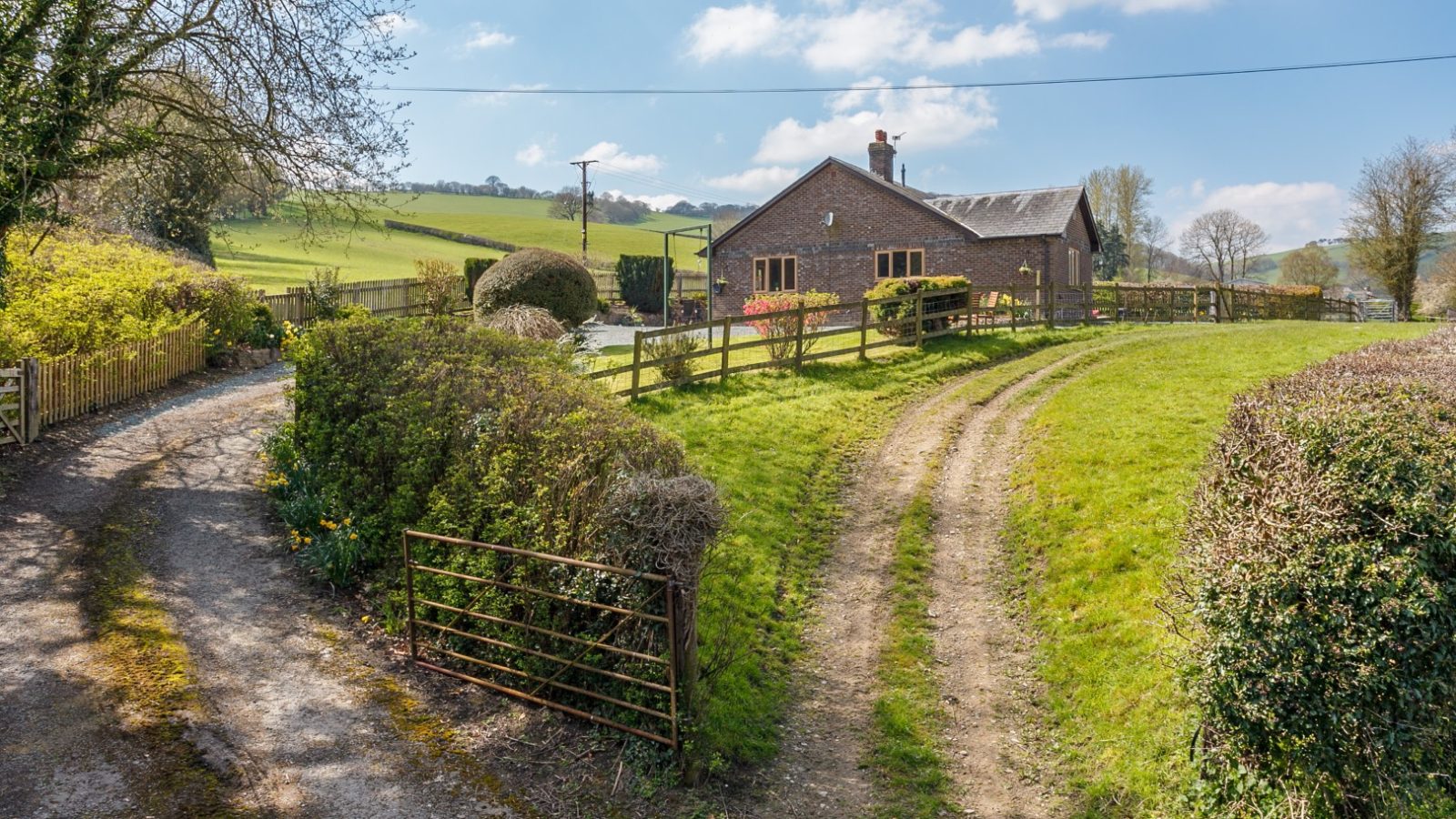 A countryside scene with Brook Cottage, winding dirt trails, lush trees, a wire fence, and a partly cloudy sky.