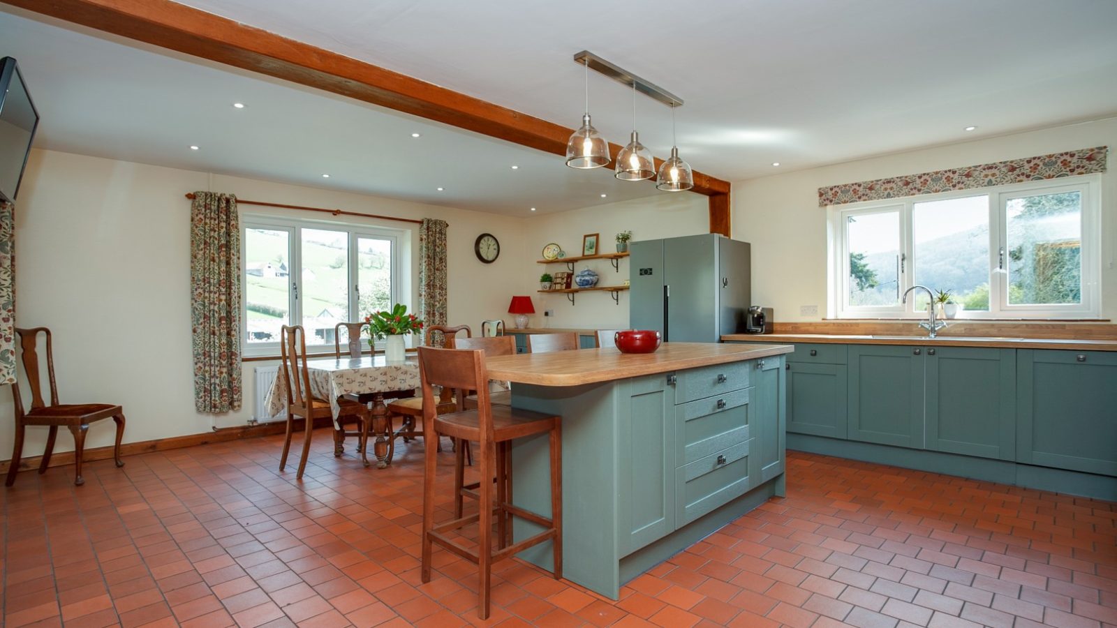 Spacious kitchen in Brook Cottage with central island, wooden chairs, green cabinets, tiled floor, and countryside views.