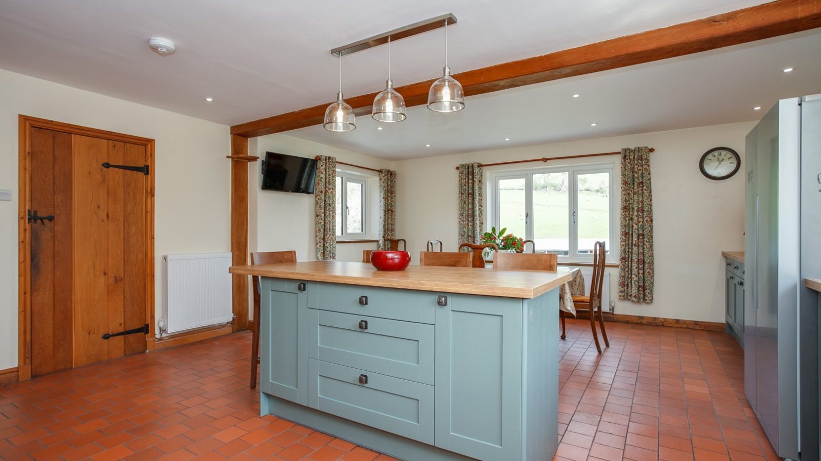 Spacious kitchen at Brook Cottage with blue island, wooden beams, pendant lights, terracotta floor, and countryside views.