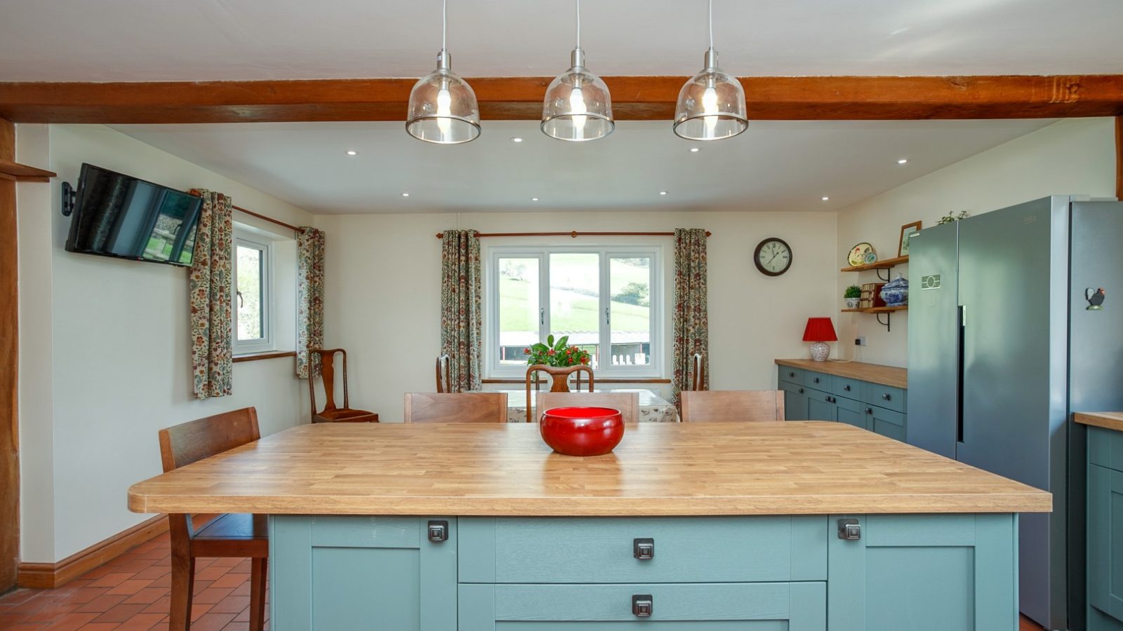 Spacious kitchen at Brook Cottage with light blue cabinets, wood island, stainless steel appliances, mounted TV, and window view.