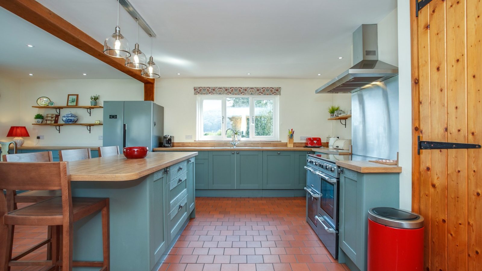 A modern kitchen in Brook Cottage with green cabinets, an island with stools, a large stove, and a red trash bin on tiled flooring.