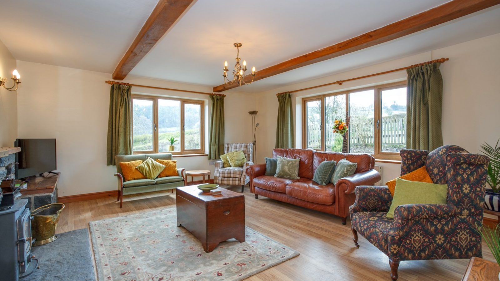 Spacious living room in Brook Cottage with wooden beams, a cozy sofa, armchairs, green curtains, and a rustic wood stove.