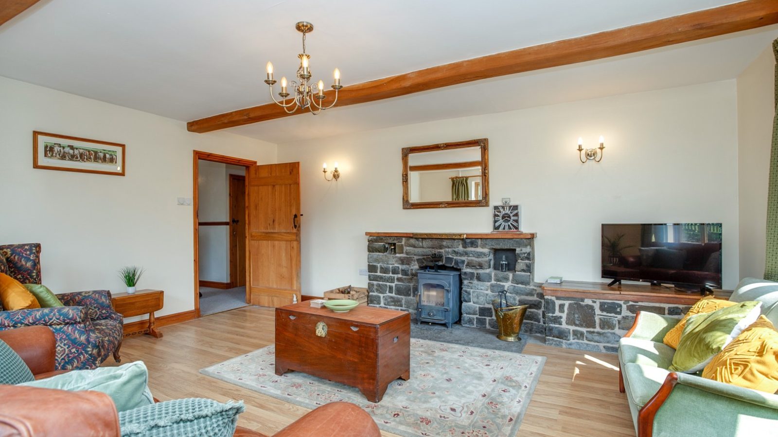 Cozy Brook Cottage living room with wooden beams, a brick fireplace, armchairs, a sofa, coffee table, and rug on wooden floors.