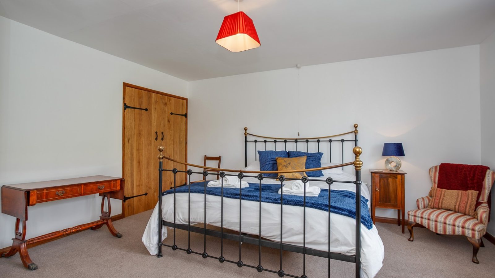 Brook Cottage bedroom with iron bed, wood furniture, striped chair, red lampshade. White walls and carpeted floor.