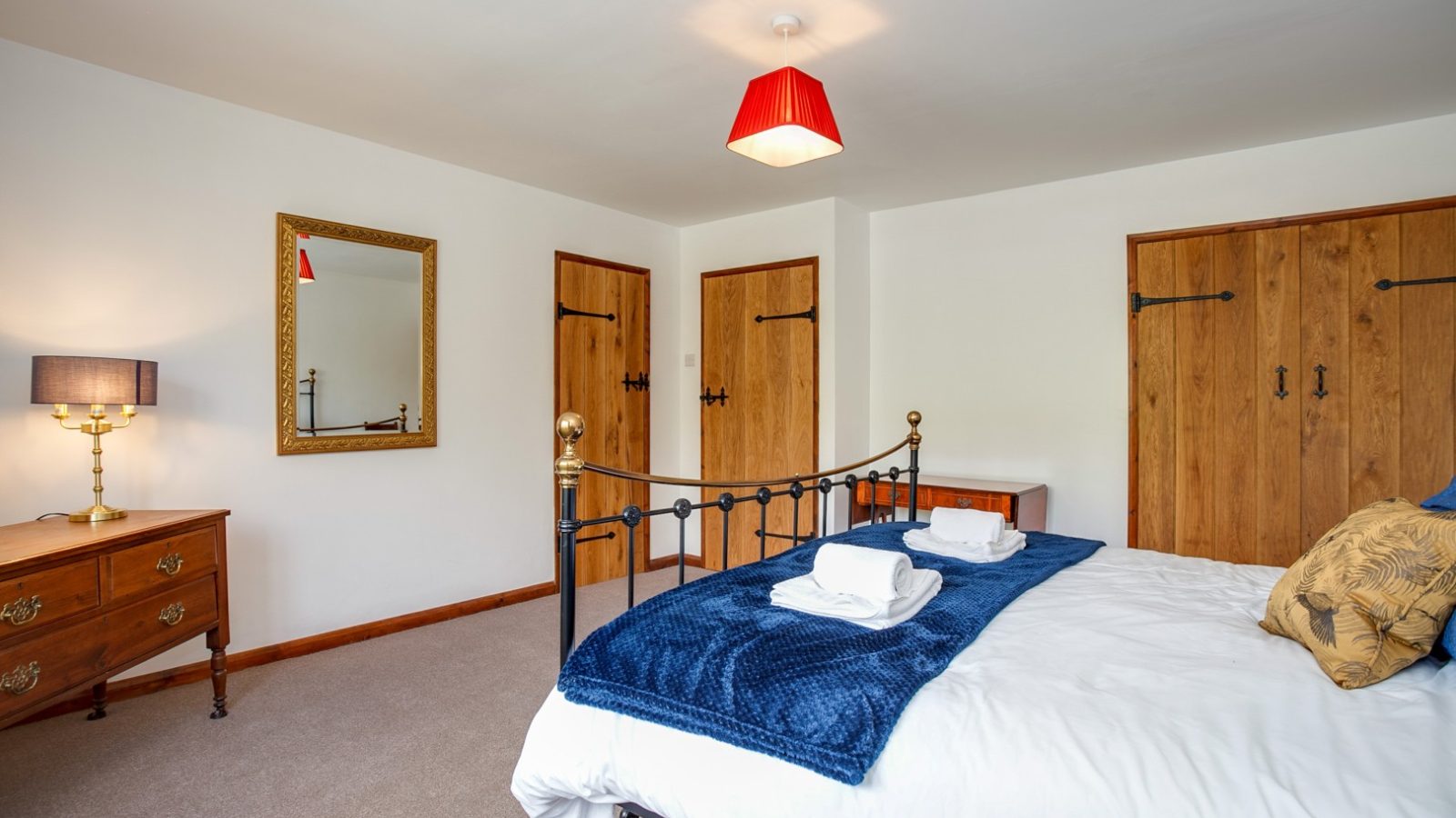 Brook Cottage bedroom: double bed, blue throw, wooden dressers, mirror, red ceiling light, and fluffy towels.