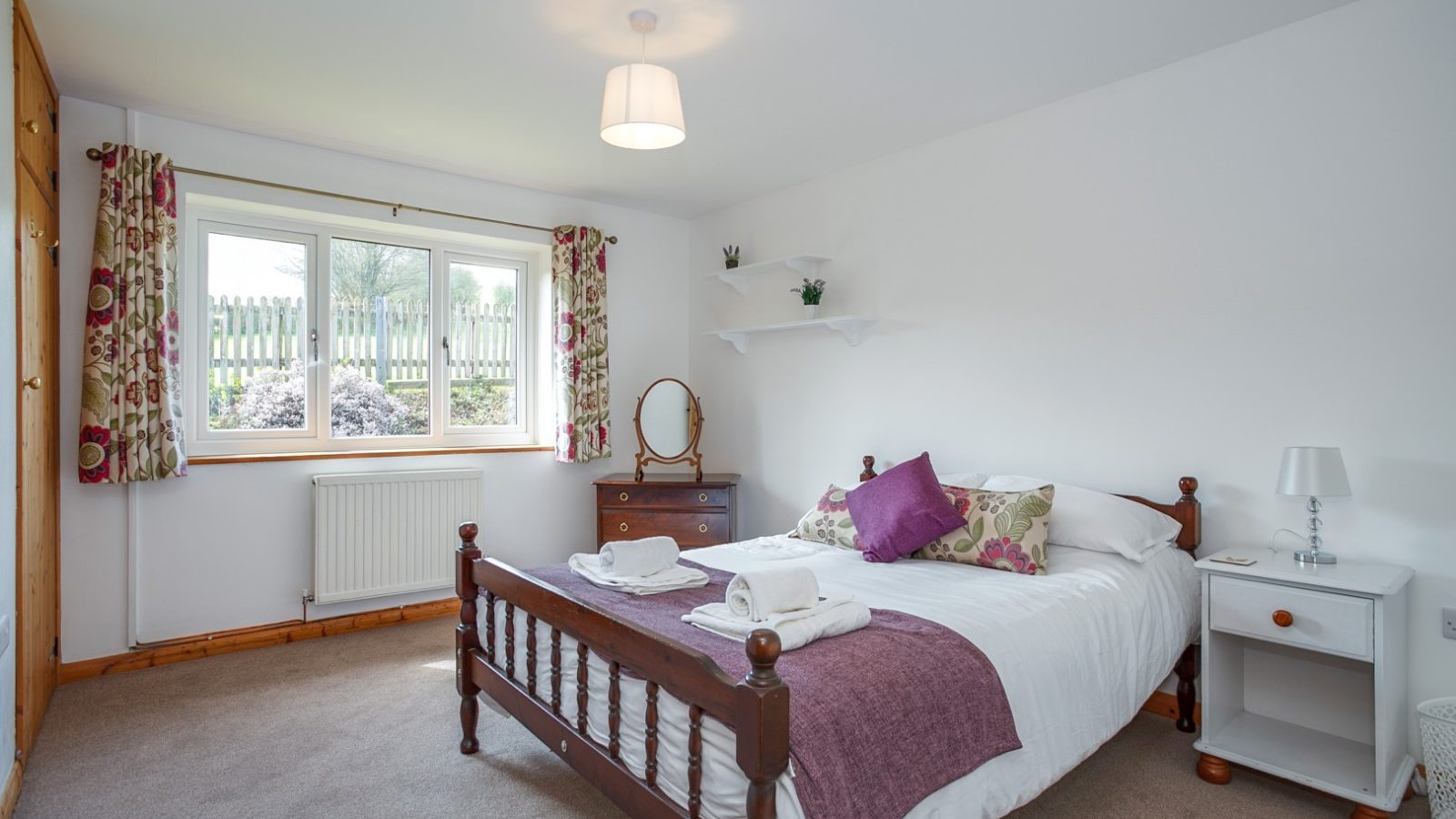 A Brook Cottage bedroom with a wooden bed, white and purple bedding, a dresser, nightstand, and floral curtained window.