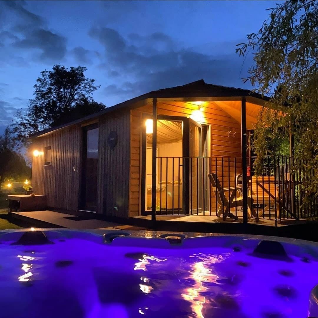 A cosy cabin with a lit porch at dusk, viewed from a bubbling hot tub with purple lights—perfect for February half-term breaks.