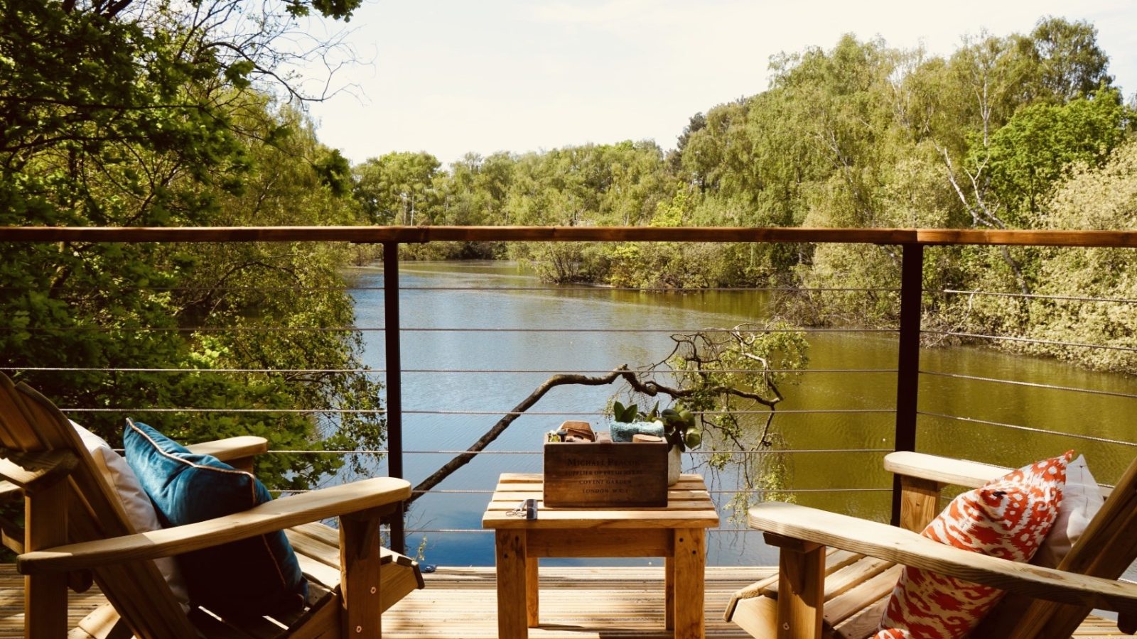 Two wooden chairs on a Treetop Hideaway deck with a small table, overlooking a serene lake surrounded by trees.