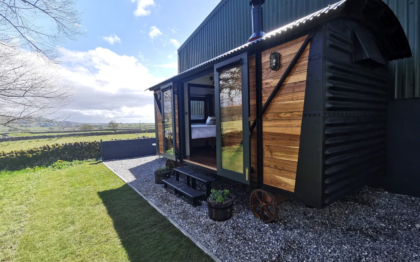 A small, rustic cabin for romantic UK getaways. With wooden siding and a metal roof sits on gravel amid the hills, under a partly cloudy sky.