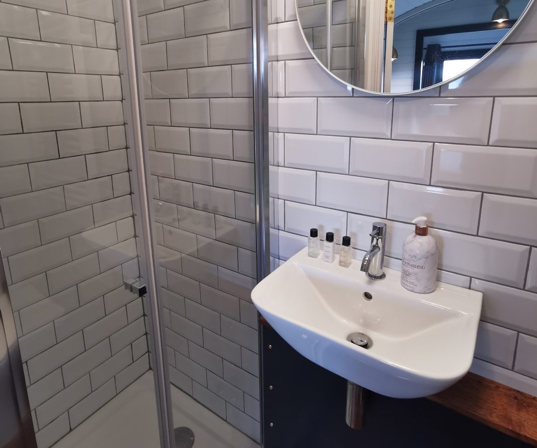 Bathroom with a glass shower, white tiles, round mirror, sink, and toiletries—mirroring the charm of Derbyshire Hills.