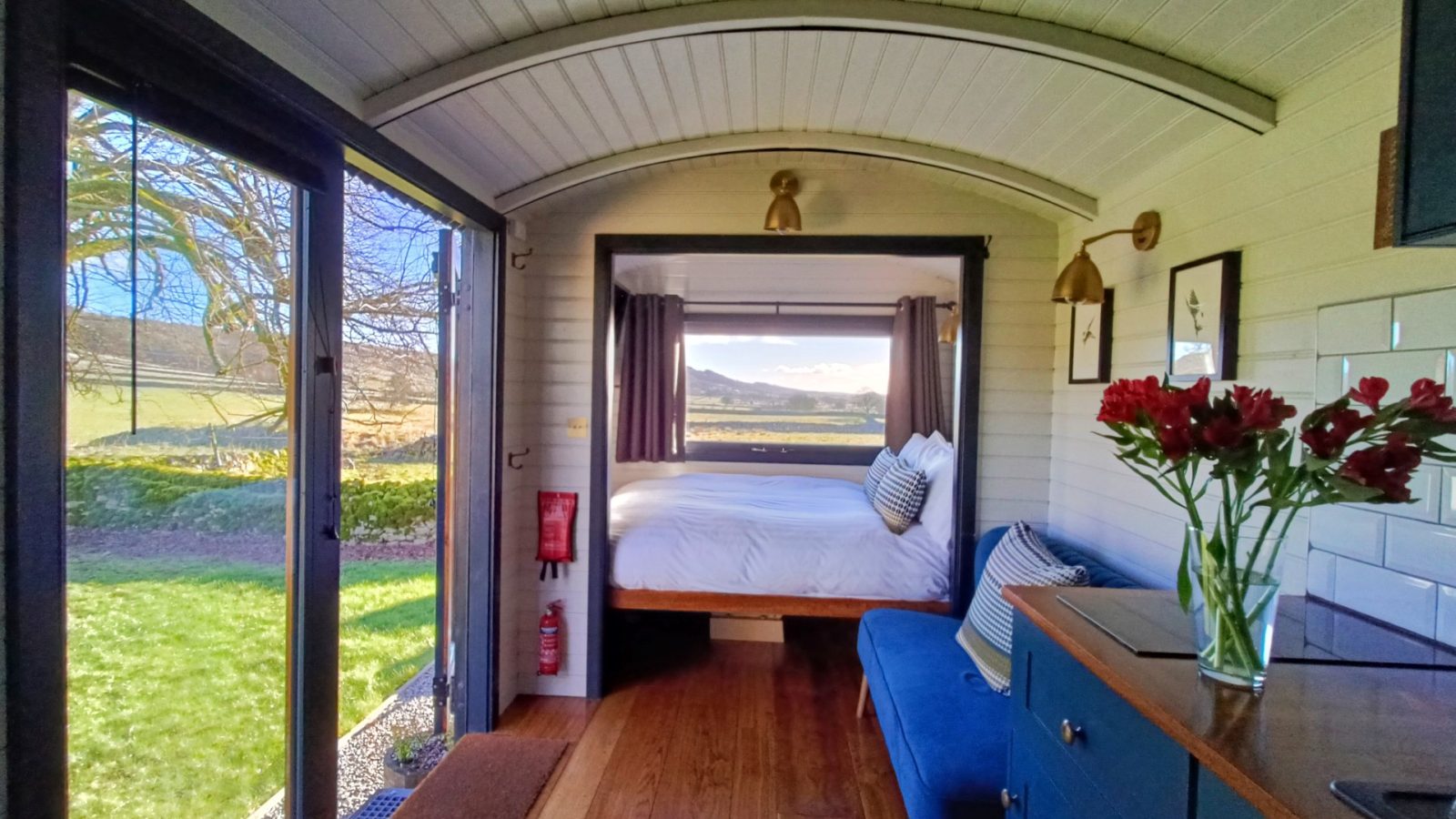 Cosy shepherds hut with homely interiors and a large bed and window view of Derbyshire Hills. Red flowers adorn the counter.