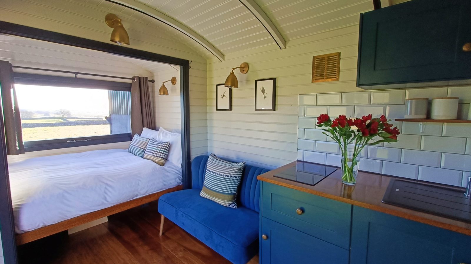 Romantic tiny home with a bed, blue sofa, and kitchen. Sunlight streams through a window with Derbyshire hills view.