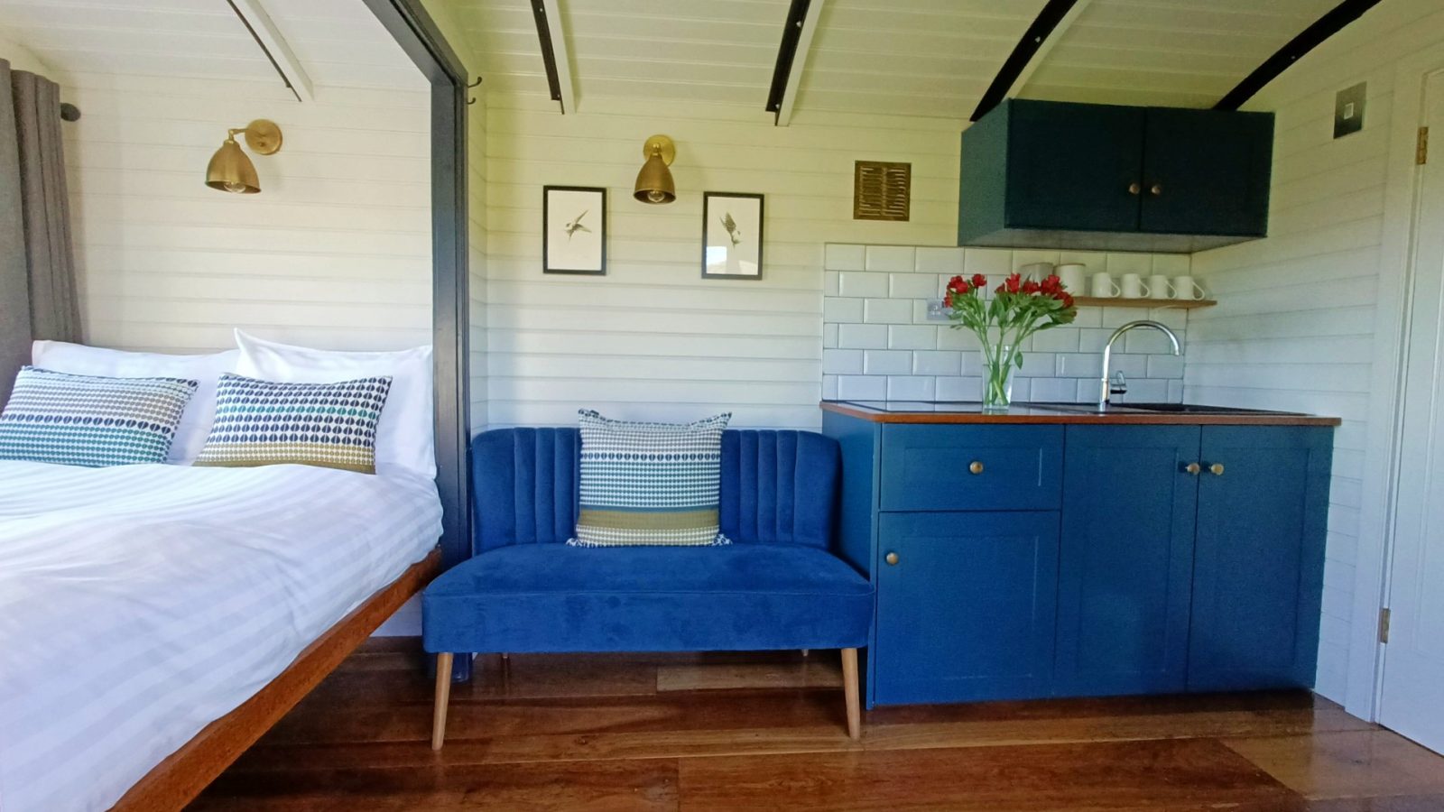 Cozy interior in a shepherd's hut with a bed, blue couch, and compact kitchenette featuring blue cabinets and fresh flowers.