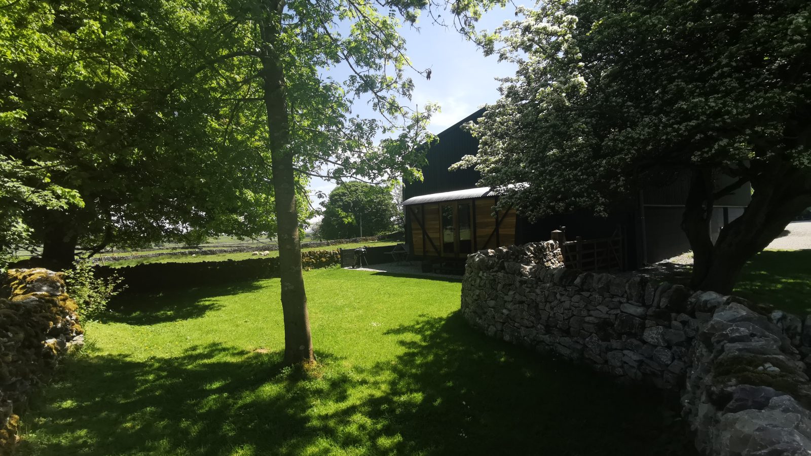 A lush green lawn with trees and a stone wall surrounds a barn in the Derbyshire Hills under a partly cloudy sky.