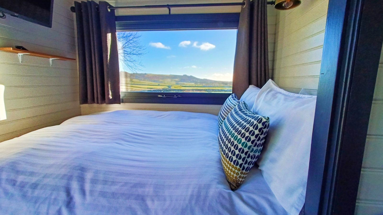 Cosy bedroom in a romantic Shepherds Hut for a couples weekend getaway. With a large window view of the sunny Derbyshire Hills, featuring a neatly made bed and polka dot pillow.