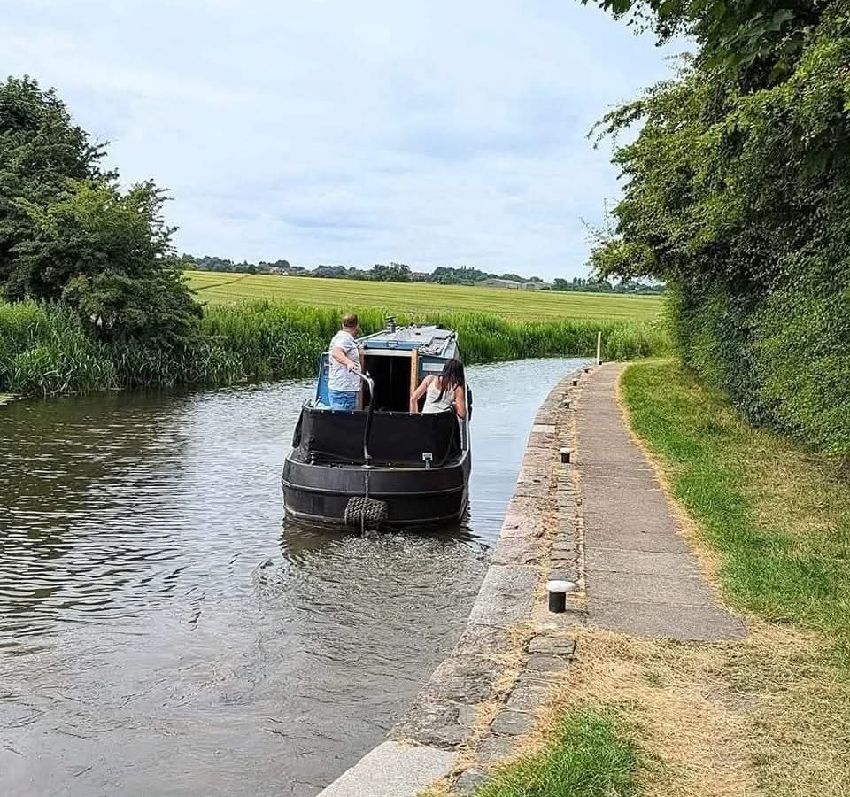 Two people navigate their narrowboat along a calm canal at Hawthorn Hideaway, with greenery and a path lining the right side.