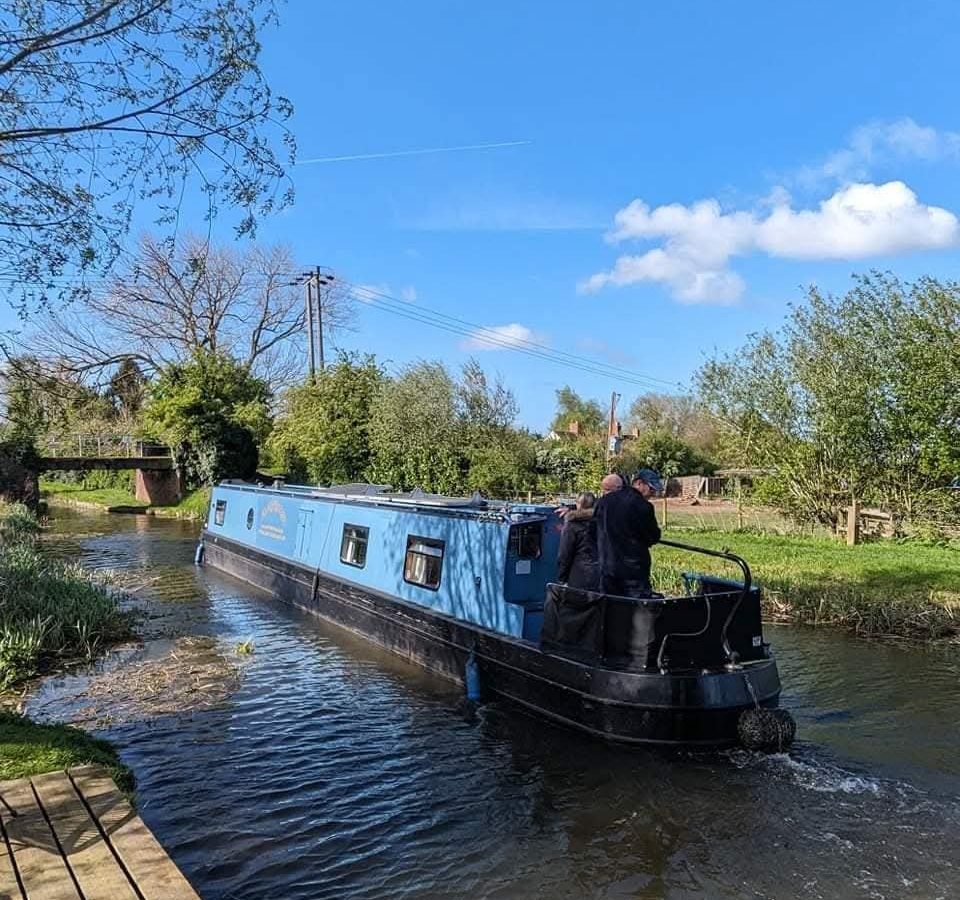 A blue narrowboat navigates a hawthorn-lined canal under a clear sky, with two people visible at the rear of the vessel.