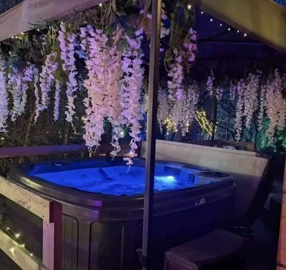 Hawthorn Hideaway: A hot tub under a canopy, adorned with hanging white flowers and string lights in a tranquil evening setting.