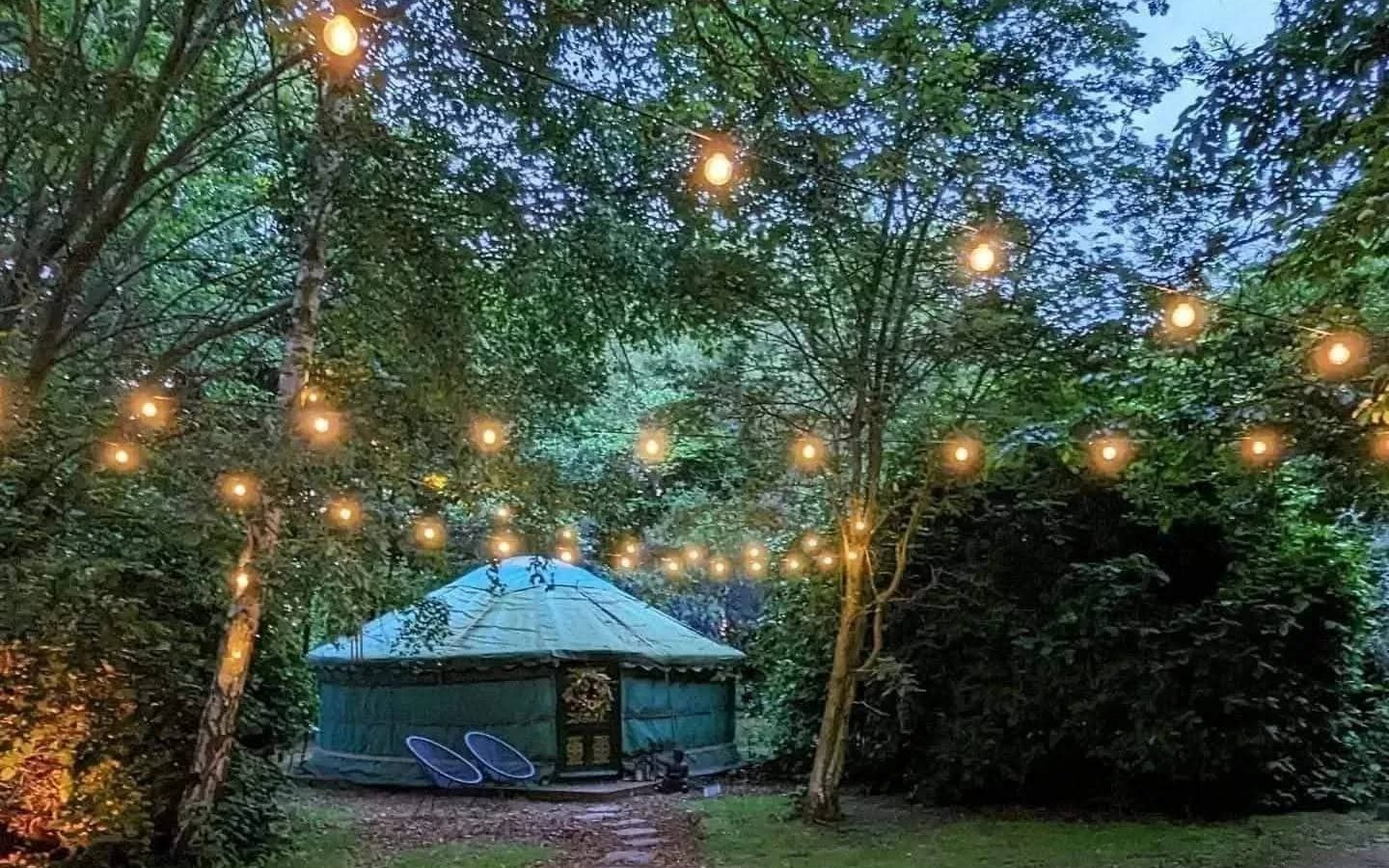 Hawthorn Hideaway: A yurt nestled among trees with string lights, set on a grassy lawn and a stone path leading to a cozy retreat.