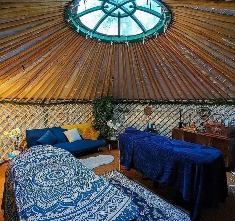 Interior of a Hawthorn Hideaway yurt with skylight, two massage tables, a blue sofa, and plants, adorned in patterned fabrics.