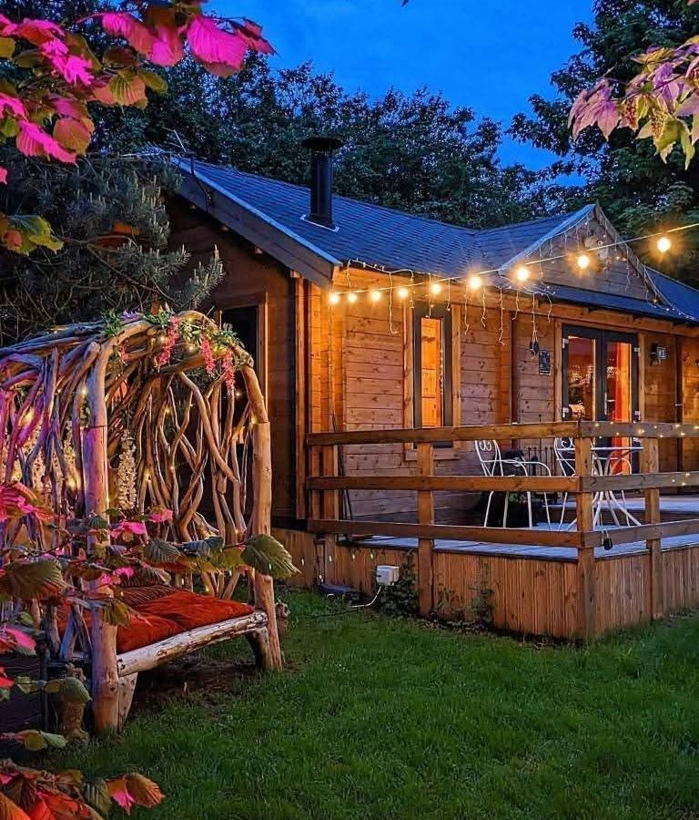 Hawthorn hideaway: A wooden cabin lit by string lights, nestled among trees with a wicker swing seat on the lawn.