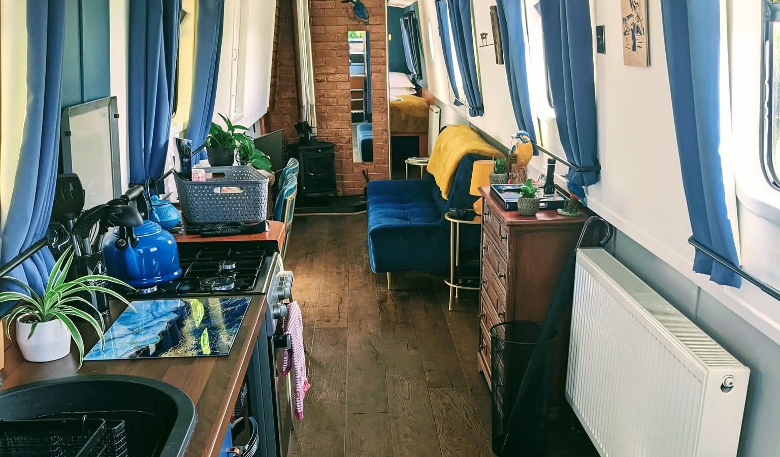 Interior of Hawthorn Hideaway, a narrowboat with a kitchen, plants, blue and yellow decor, wooden flooring, and large windows.