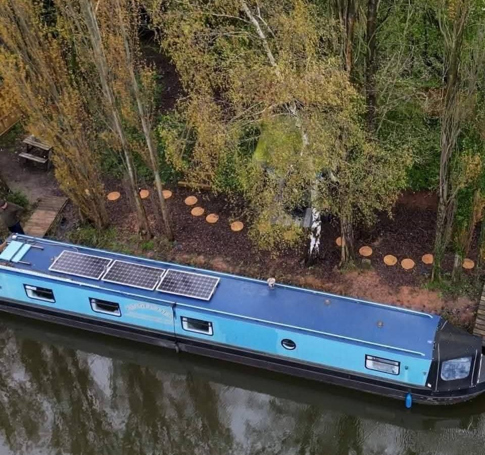 A blue narrowboat with solar panels, named Hawthorn Hideaway, is moored along a tree-lined canal with a small path nearby.