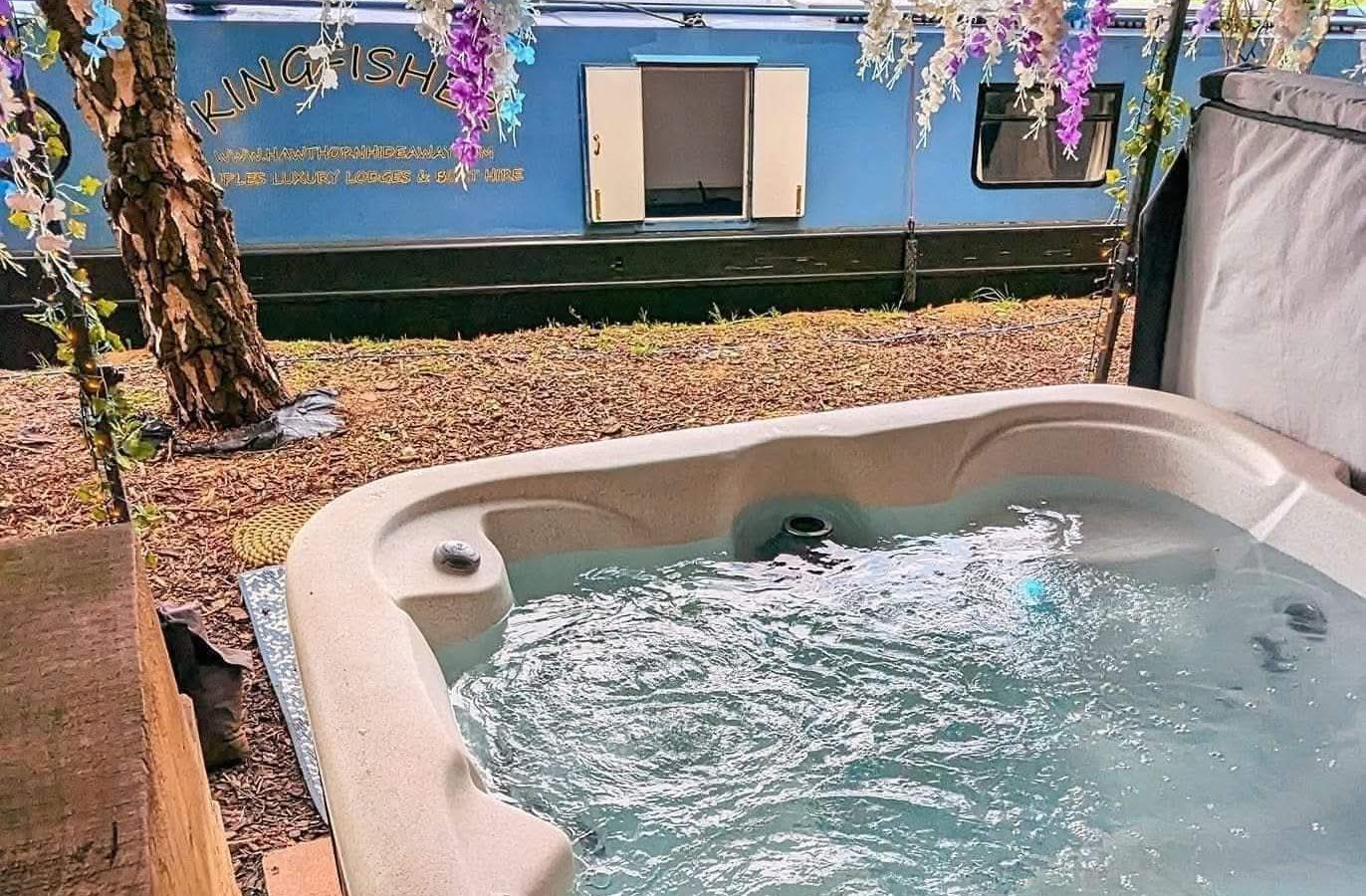 Hawthorn Hideaway features an outdoor hot tub under hanging flowers, with a blue canal boat in the background.