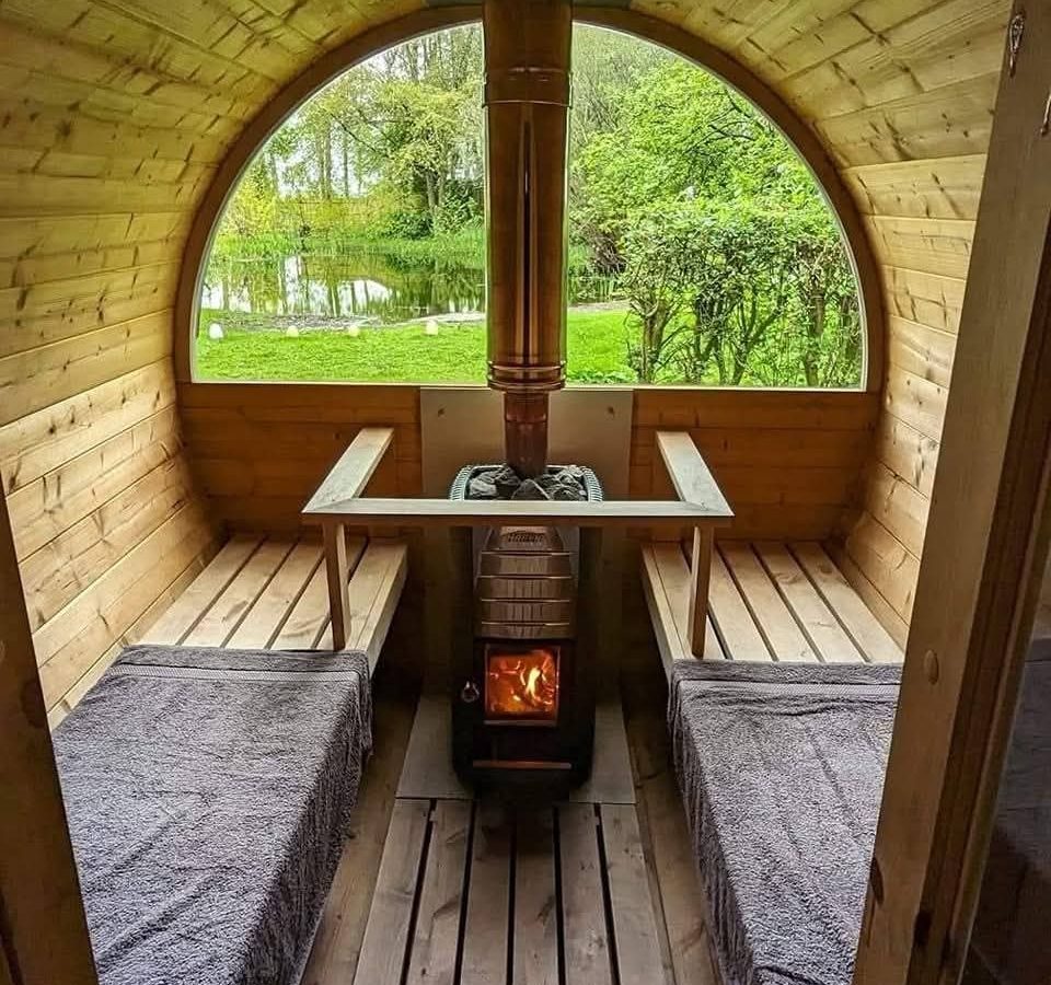 Interior of Hawthorn Hideaway sauna with a central stove, benches, and a view of greenery through a large window.