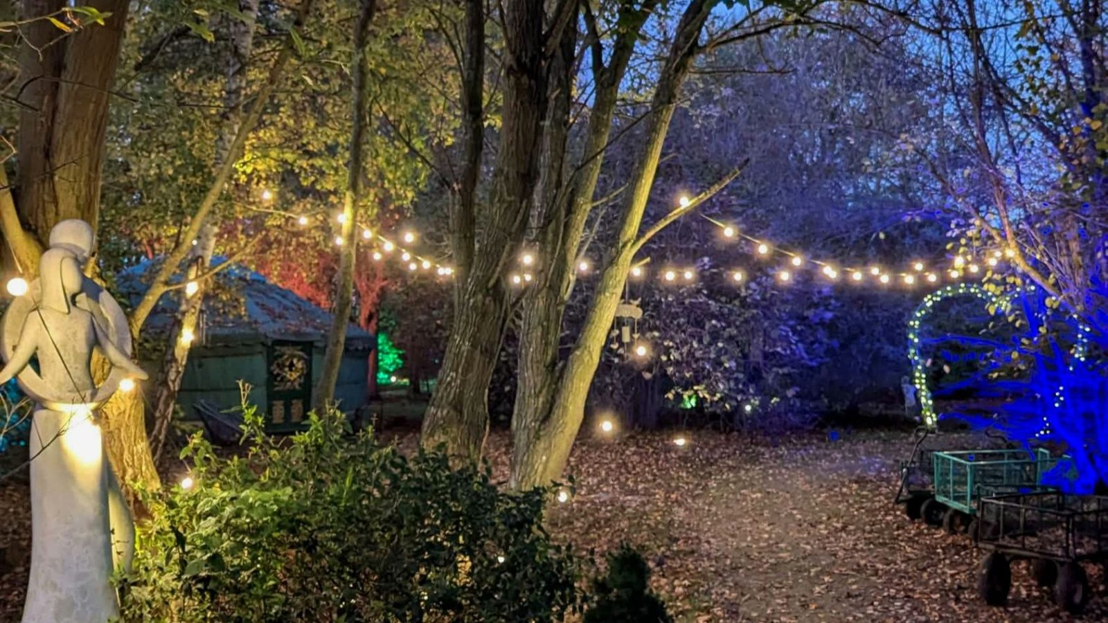 Hawthorn Hideaway: A garden pathway at dusk with trees, string lights, a statue, and a small cart on the right.