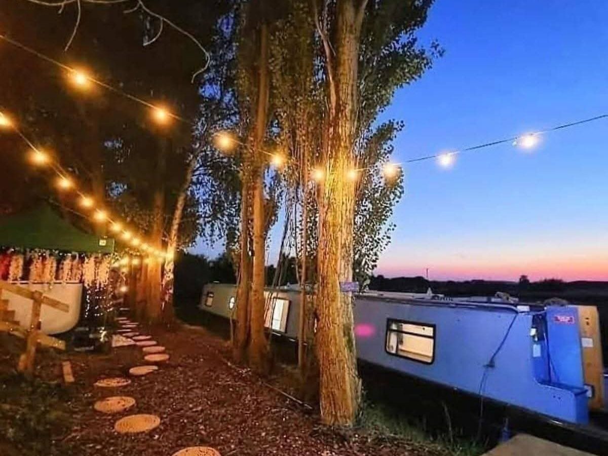 Path with round stepping stones and string lights beside a blue houseboat, a twilight hideaway under the hawthorn sky.