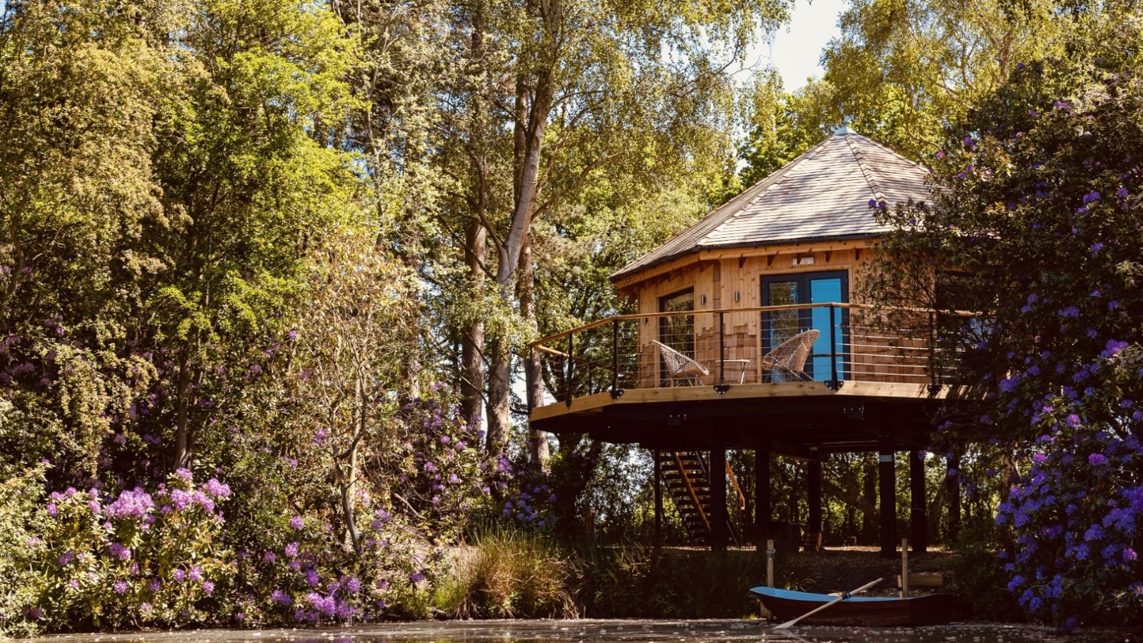 Treetop hideaway on stilts amid lush greenery and purple flowers, overlooking a calm pond with a small boat in the foreground.